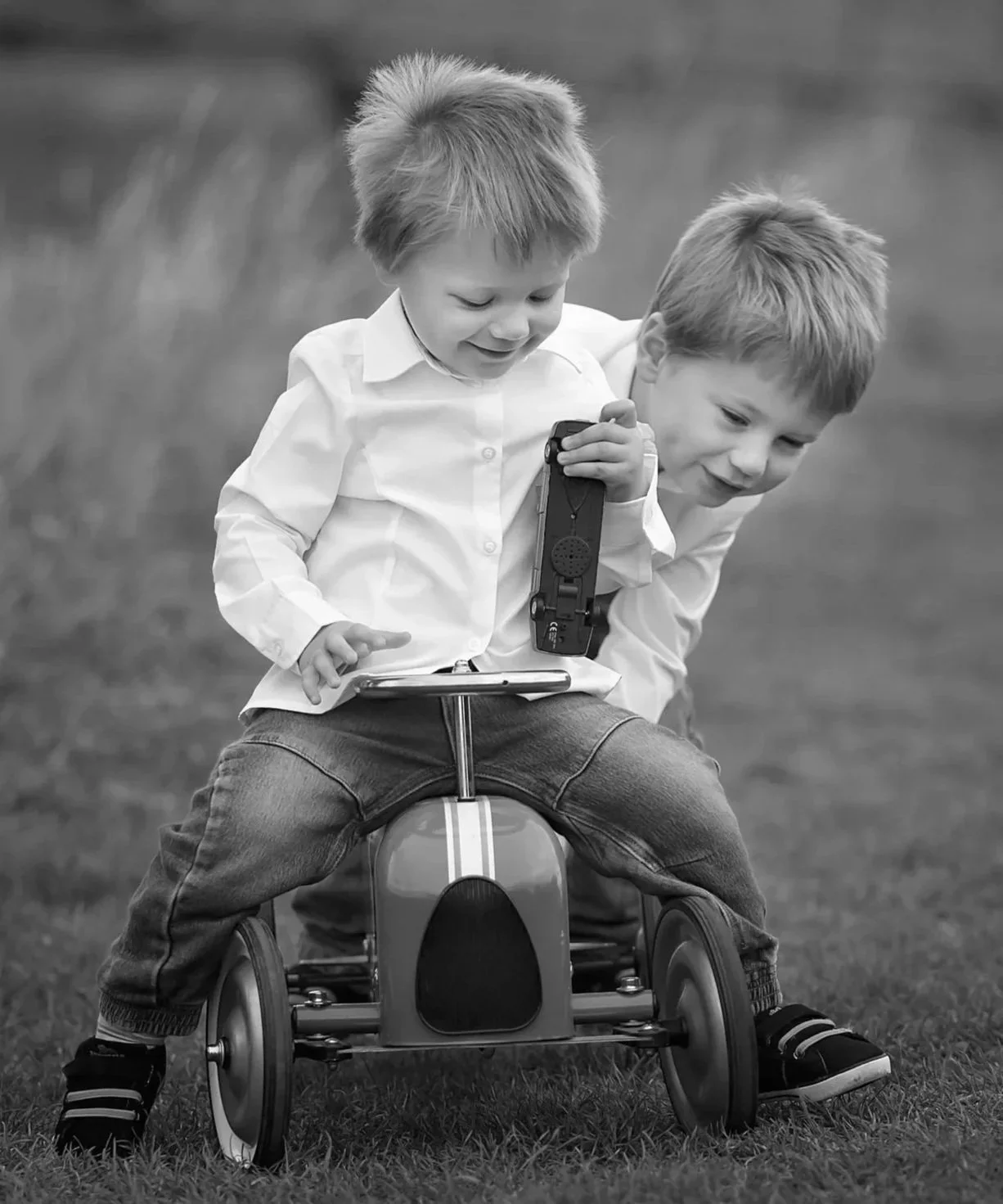 Two young boys outdoors, one sitting on a ride-on toy car and holding a tablet, the other leaning close, both smiling and looking at the tablet.