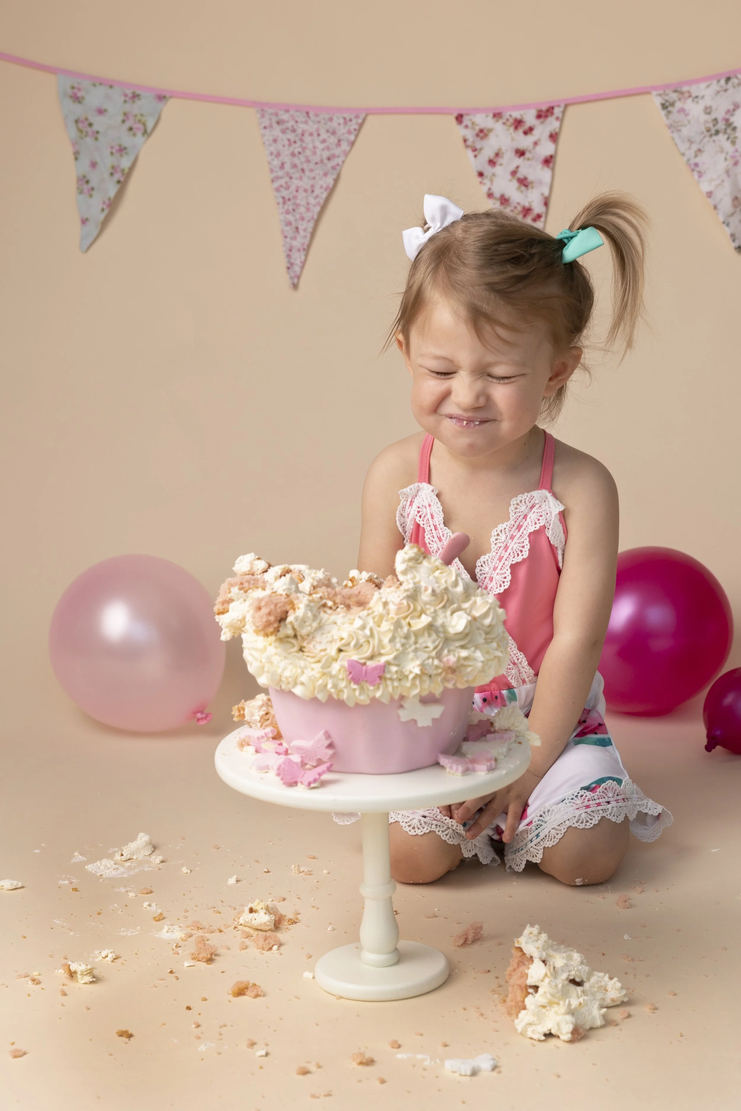 Young girl with pigtails and a white bow, wearing pink and white lace dress, kneeling behind a pink cake with whipped cream and pink decorations, at a cupcake-themed party, with pink and purple balloons and bunting in the background, crumbs and cake pieces scattered around.