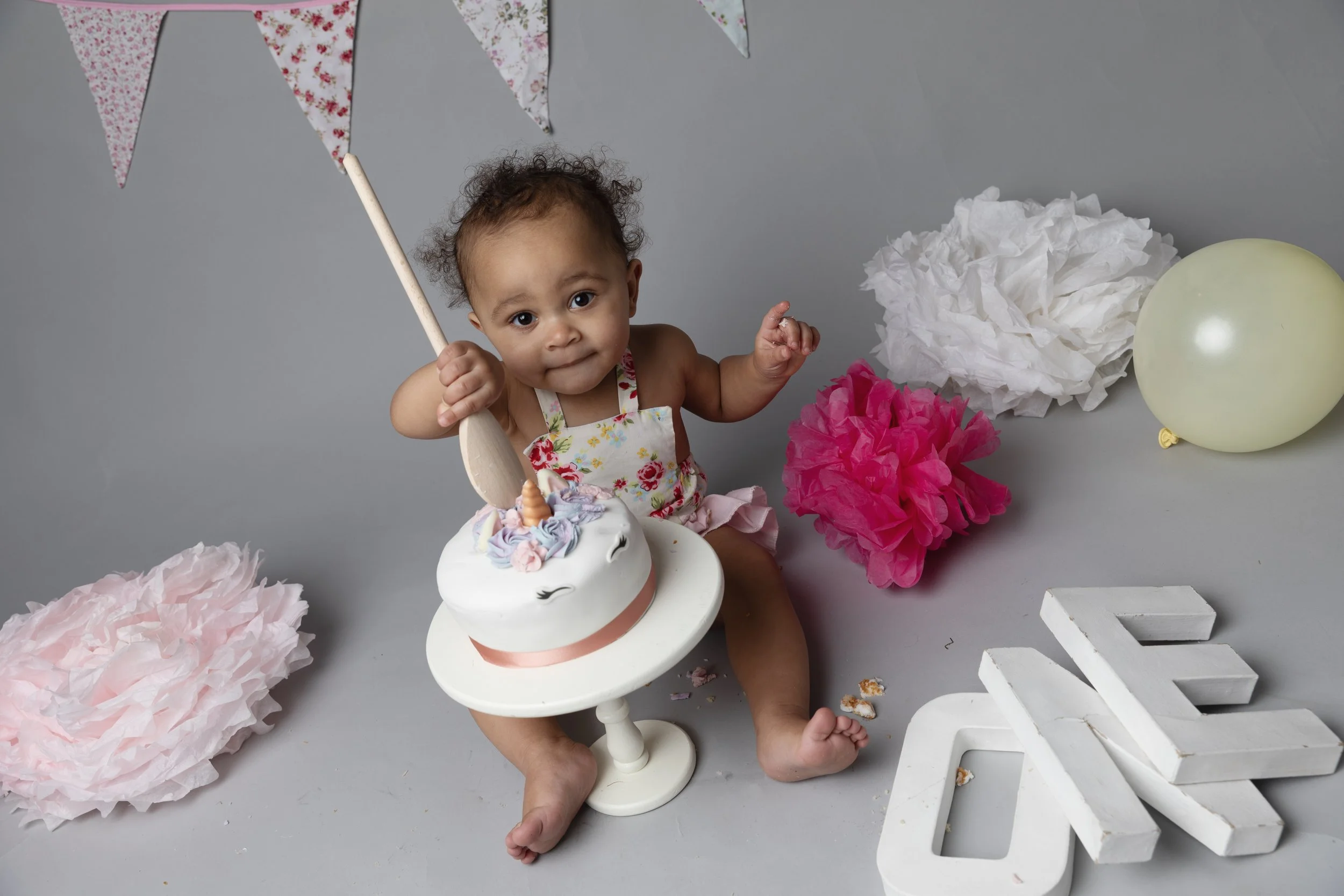A young child celebrating a birthday with a unicorn-themed cake surrounded by pink and white paper decorations in a festive setting.