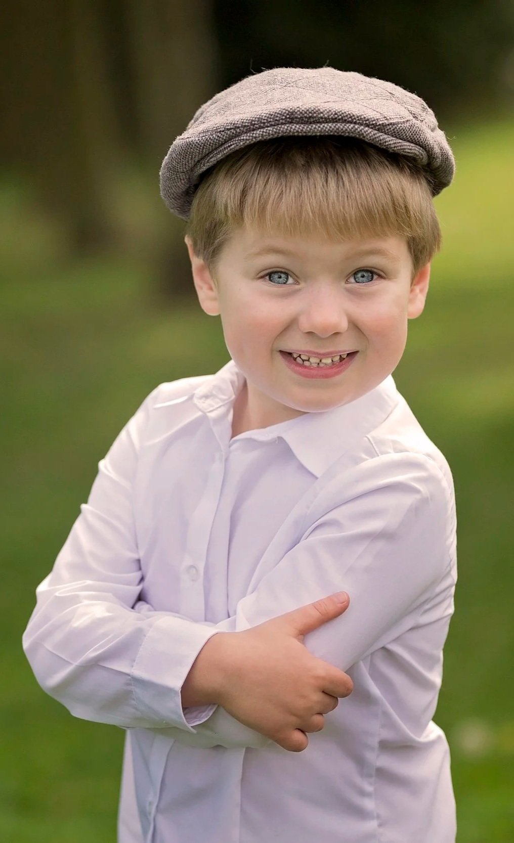 A young boy with blond hair and blue eyes, smiling, wearing a white shirt and a gray flat cap, standing outdoors with a blurred green background.