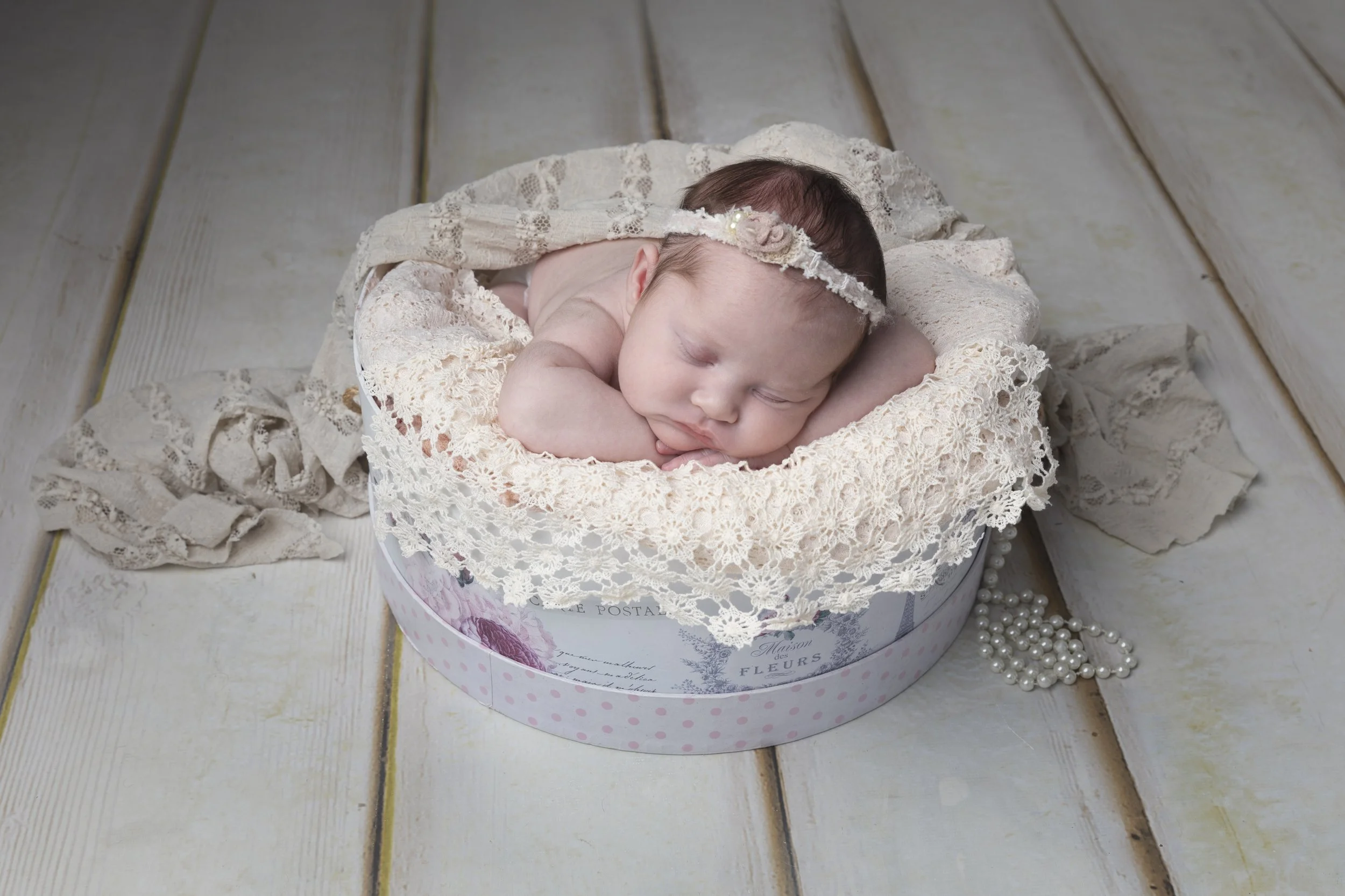 A sleeping baby in a vintage lace bonnet, lying on a lace blanket inside a round floral box, with a string of pearls beside it, on a wooden surface.