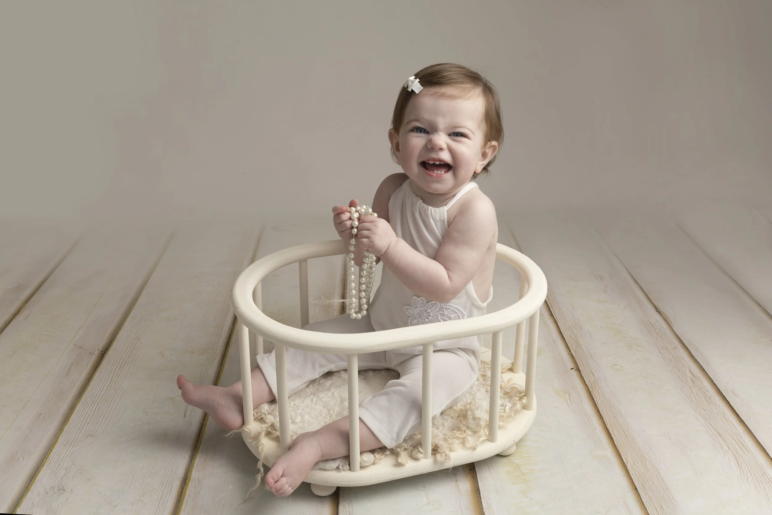 A baby girl sitting in a white circular basket on a wooden floor, holding pearl necklaces, making a playful face with scrunched nose and open mouth, wearing a sleeveless white outfit with lace detailing and a white hair clip.