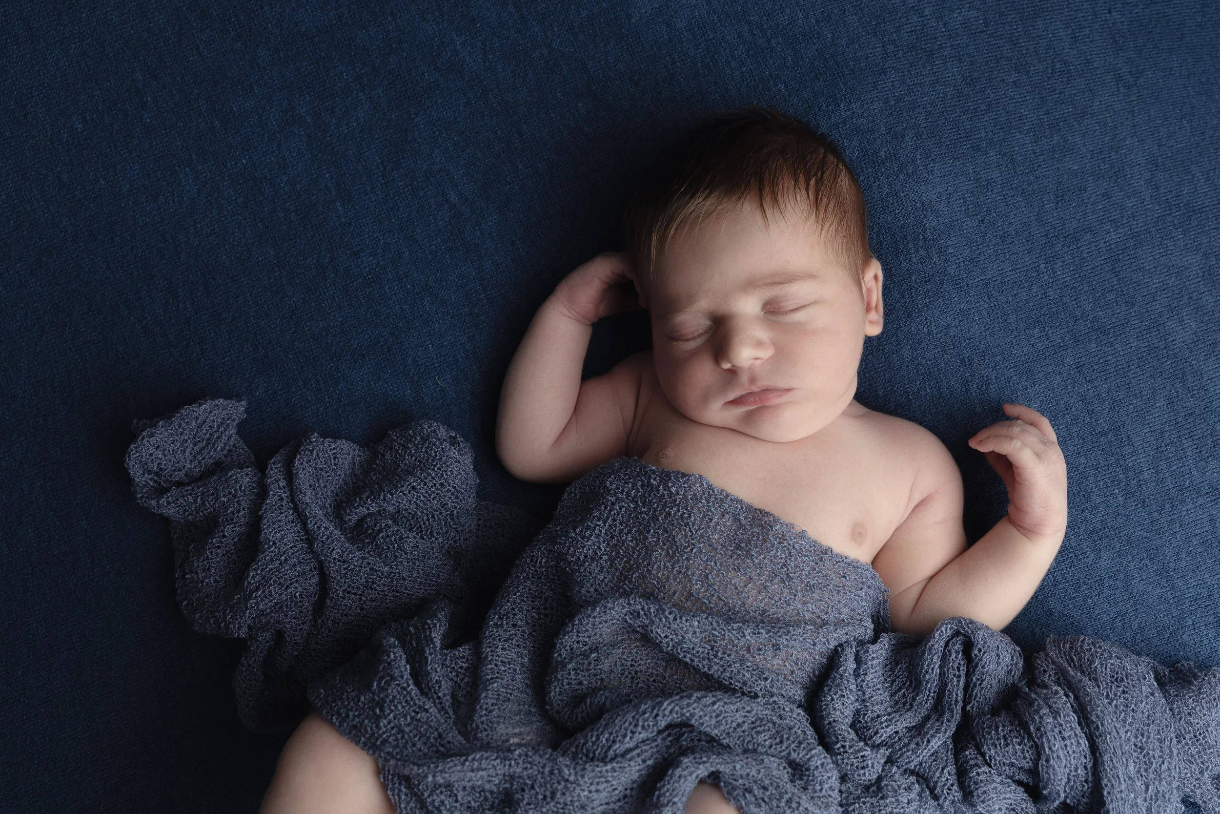 A sleeping baby with reddish hair lying on a dark blue textured surface, partially covered with a gray fabric.