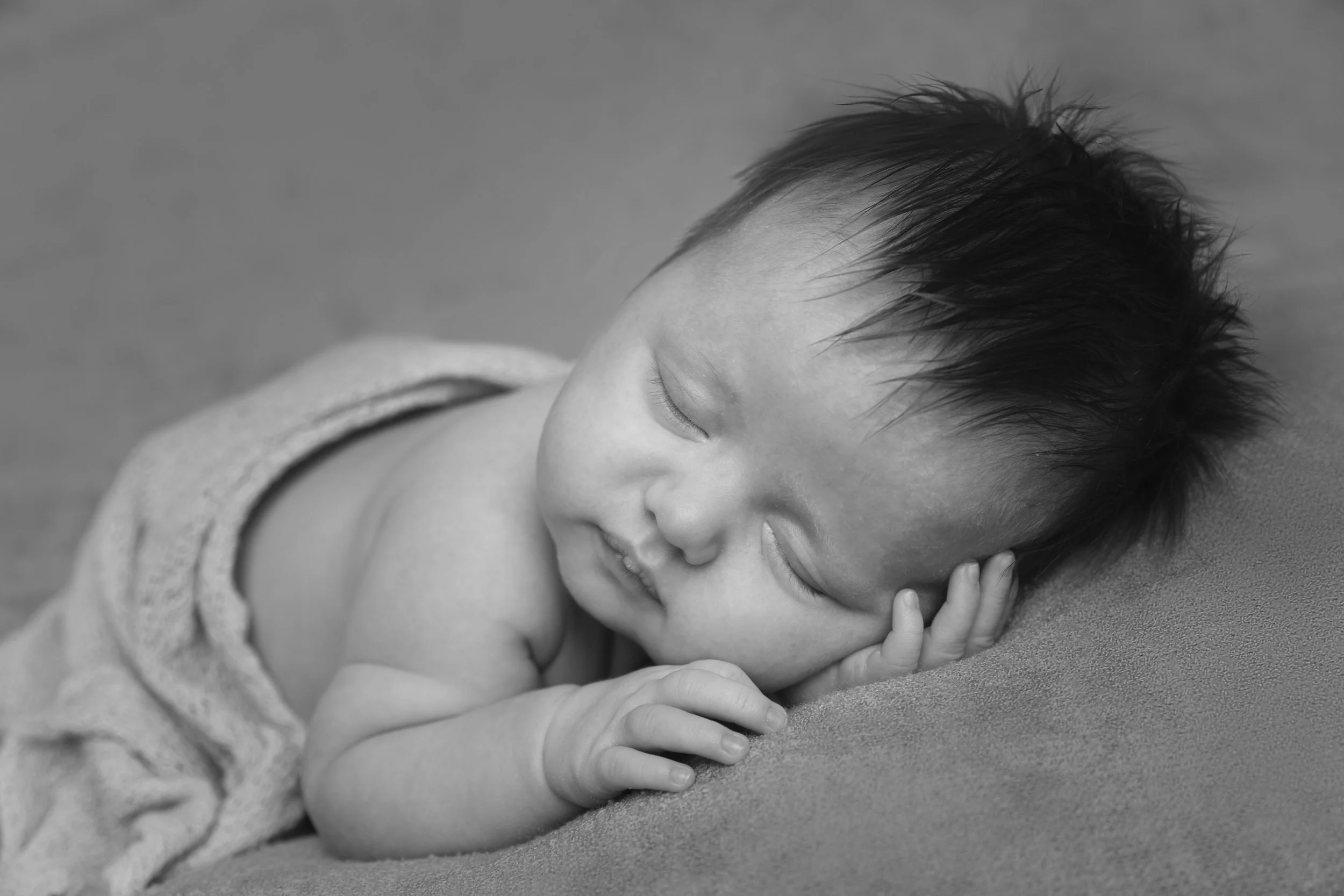 A black and white photograph of a young boy with short, spiky hair, lying on a sandy surface with his head resting on his hand and smiling gently.