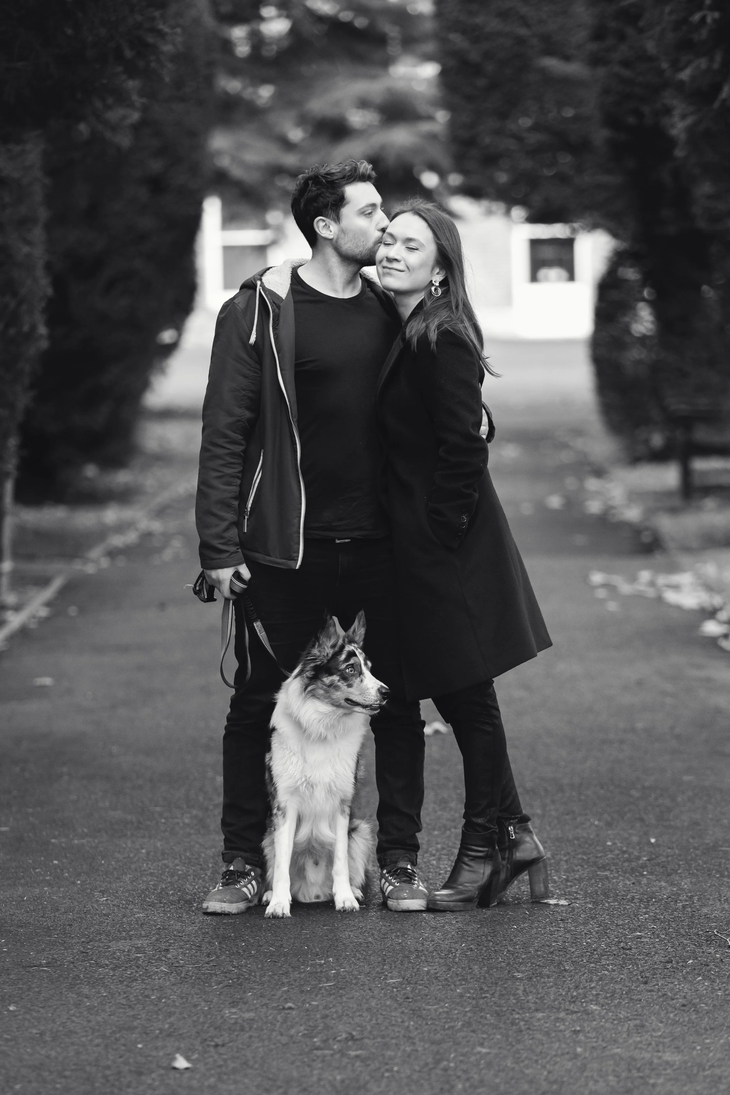 A black and white photo of a young couple with a dog standing between them on a park path, surrounded by trees.