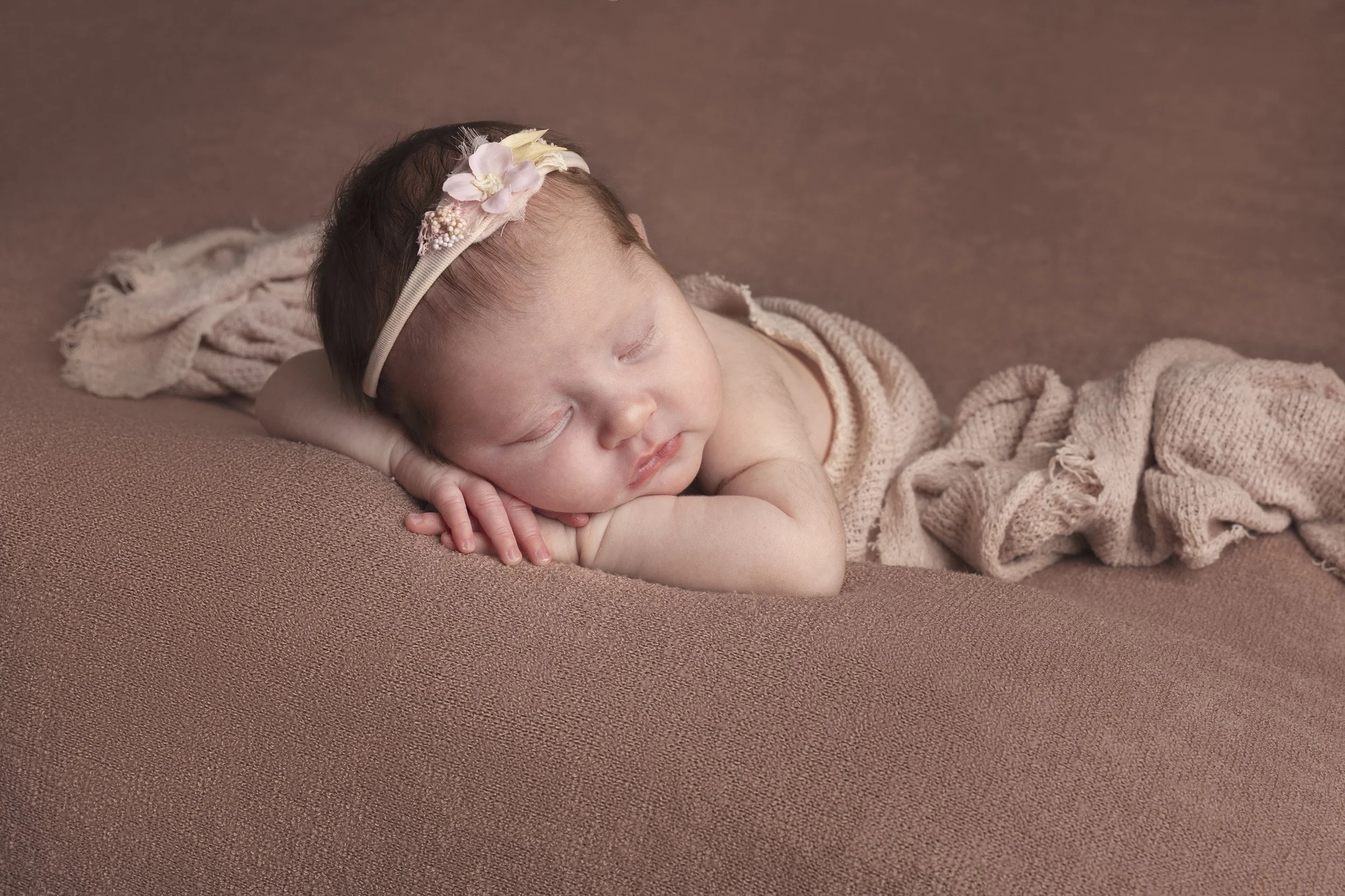 A sleeping baby lying on a pink surface, wearing a beige headband with pink flowers and a beige knitted blanket.
