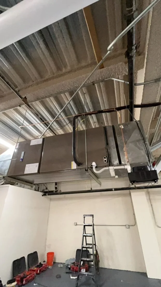 Industrial air conditioning or ventilation unit installed on a warehouse ceiling, with tools and a ladder in the room below.