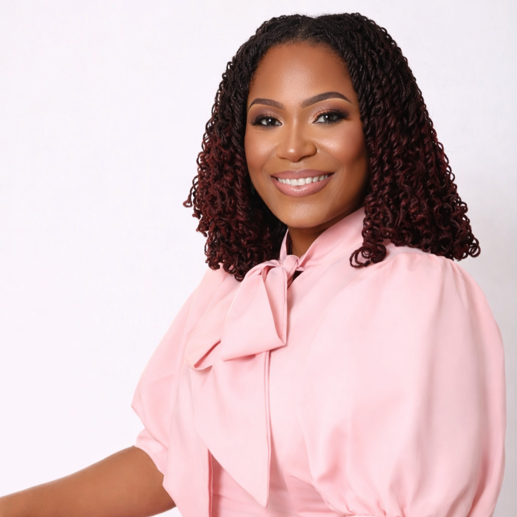 A smiling woman with curly hair wearing a light pink blouse with a bow, standing against a white background.