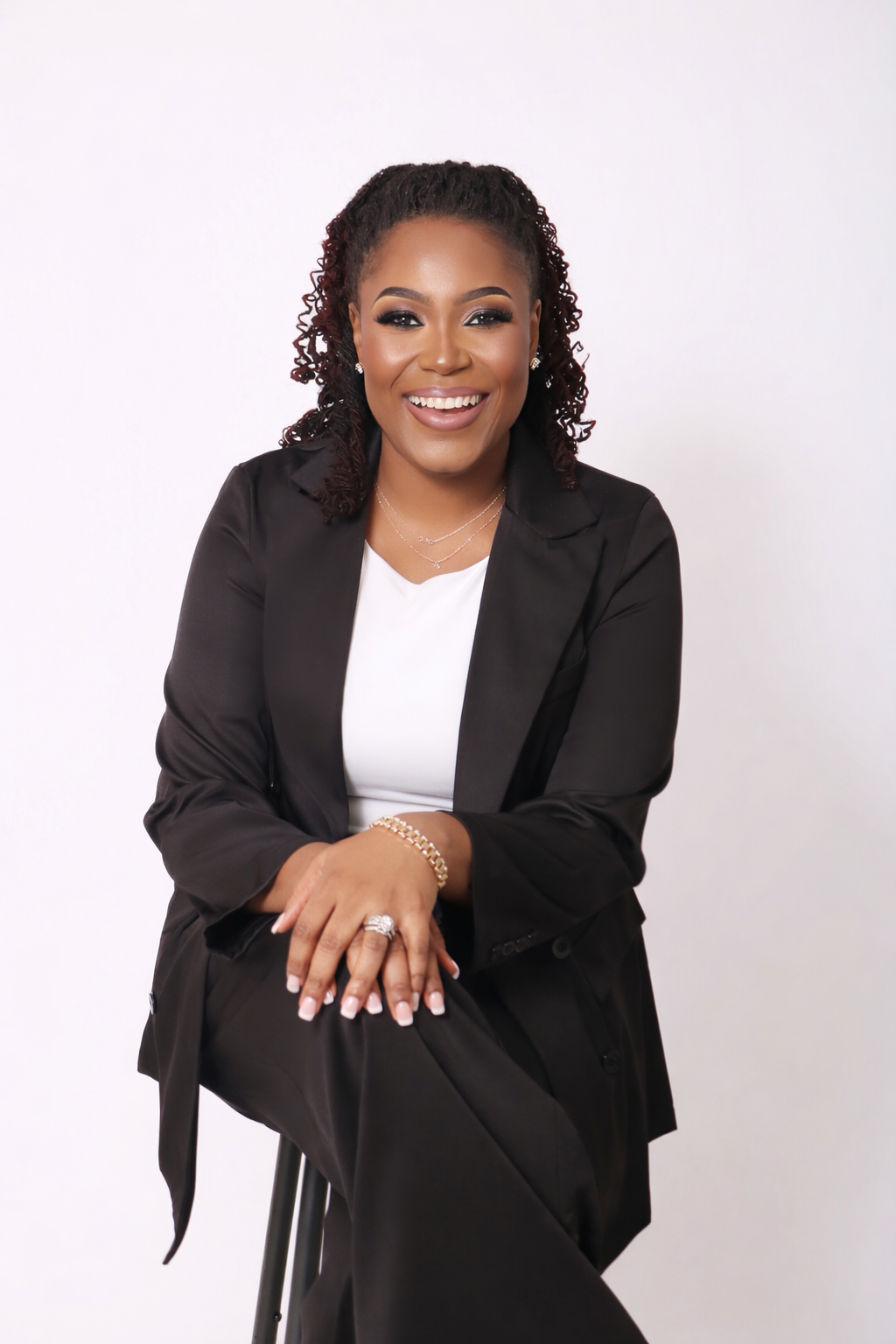 A woman with curly hair smiling, wearing a black blazer and white top, sitting on a stool with a white background.