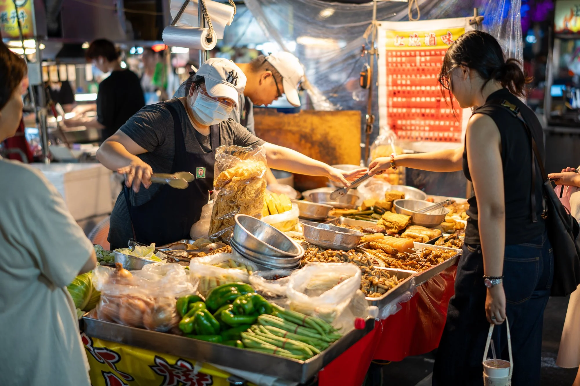 Vrouw met gezichtsmasker en pet koopt voedsel bij een streetfoodkraam met veel verschillende gerechten en groenten.