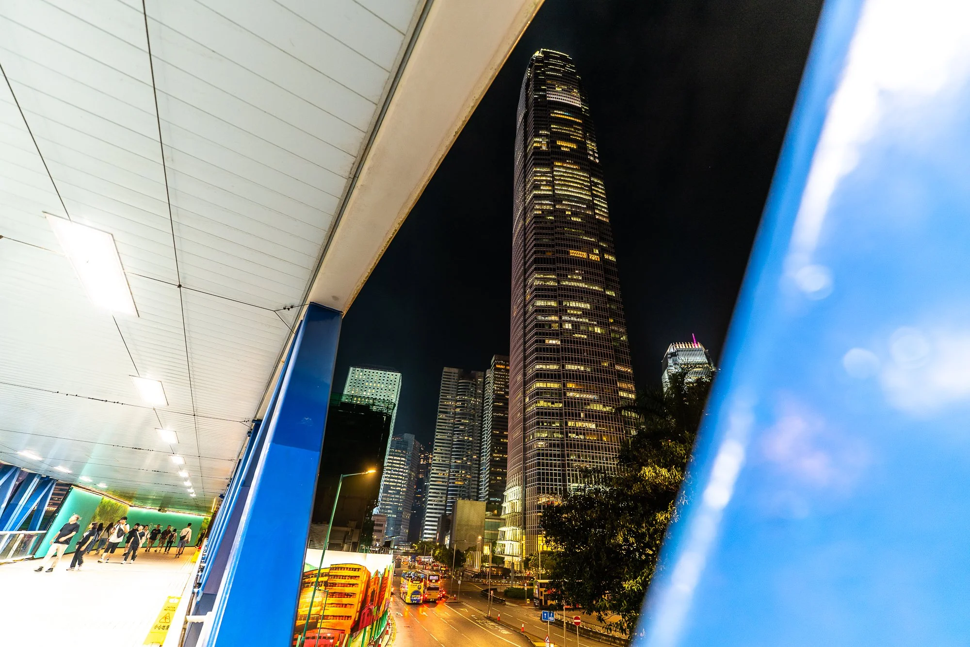 Een foto van een stad 's nachts met een hoge toren die verlicht is, een overwegend zwart hemel, een brug of structuur met blauwe details en mensen die door een ondergrondse tunnel of pas gaan.