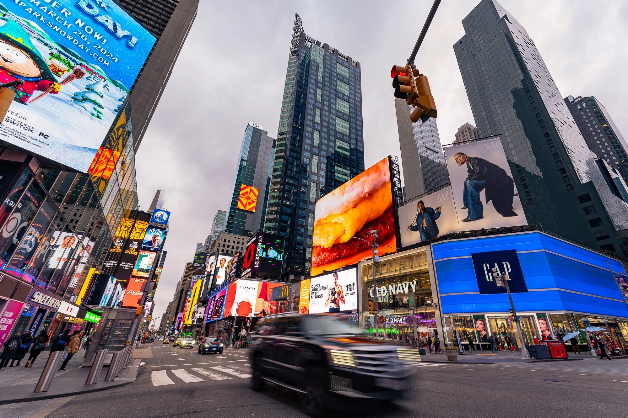 Het is een drukkere foto van Times Square in New York met digitale reclameborden, hoge gebouwen en een verkeerslicht. Er rijden auto's en er lopen voetgangers op straat onder een bewolkte hemel.