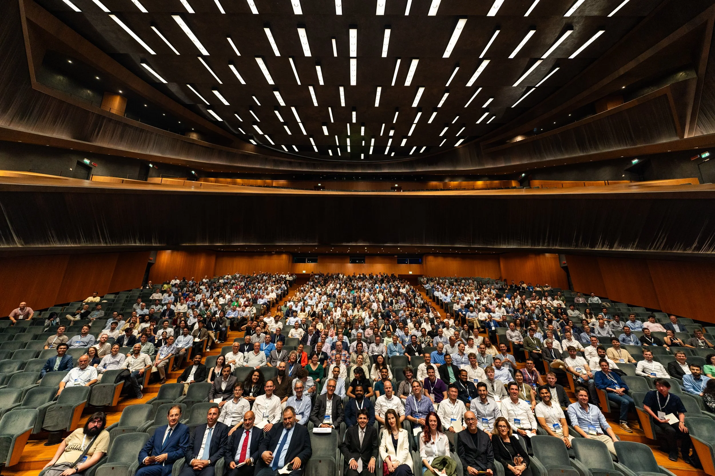 Groep mensen zit in een grote concertzaal of auditorium, klaar voor een evenement of conferentie.