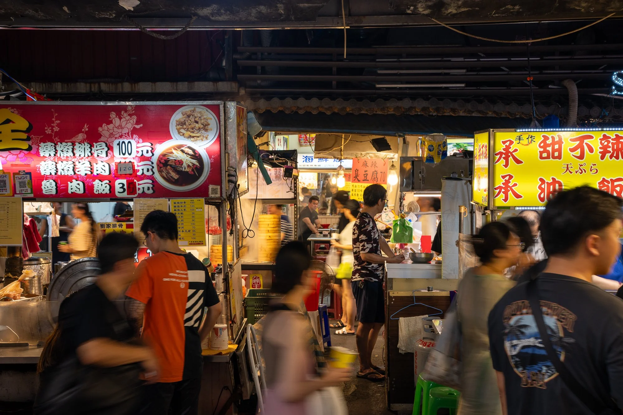 Een drukke markt met verschillende foodkraampjes, mensen die bestellen en eten, en kleurrijke borden en banners met Chinese tekens.
