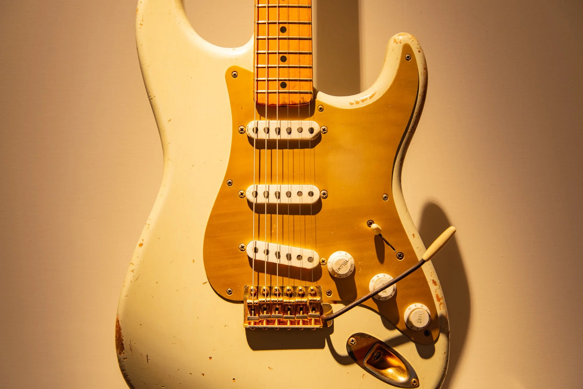 Een verouderde witte elektrische gitaar met een gouden pikkuur en verroeste onderdelen, ligt op de vloer met een plectrum ernaast.