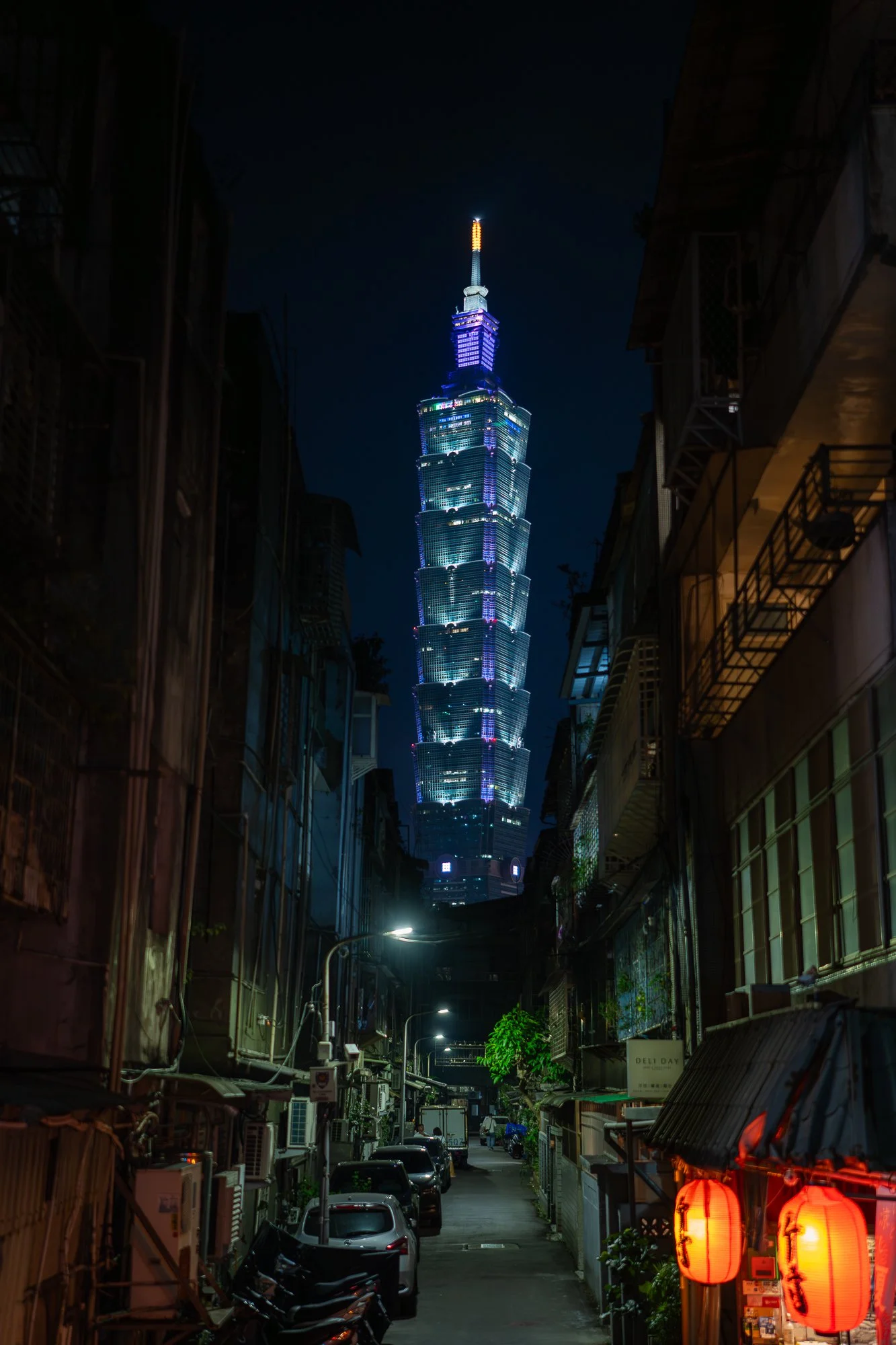 Een smalle straat met voertuigen en winkeltjes, met op de achtergrond de Taipei 101 toren verlicht in neonkleuren, 's nachts.