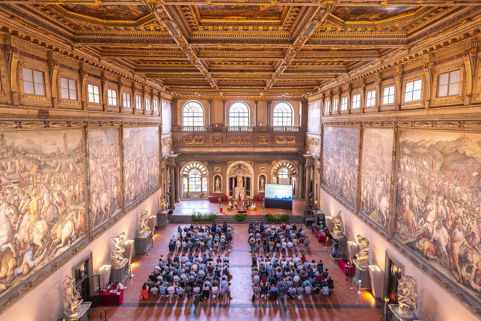 Grote zaal met hoog plafond, versieringen, grote ramen, wandkunst en een publiek dat op een podium zit voor een presentatie of ceremonie.