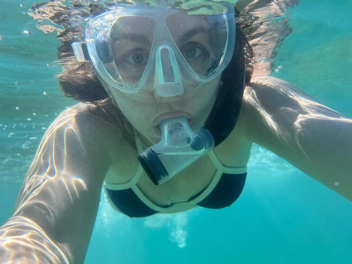 A woman snorkeling underwater, wearing a mask and snorkel, looking directly at the camera.