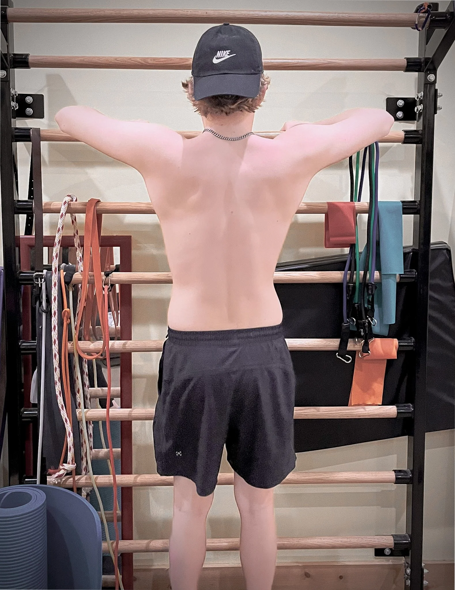 Person standing in front of a wooden gymnastic wall bar, wearing black shorts, a chain necklace, and a dark Nike cap, with arms extended and holding onto the top bar.