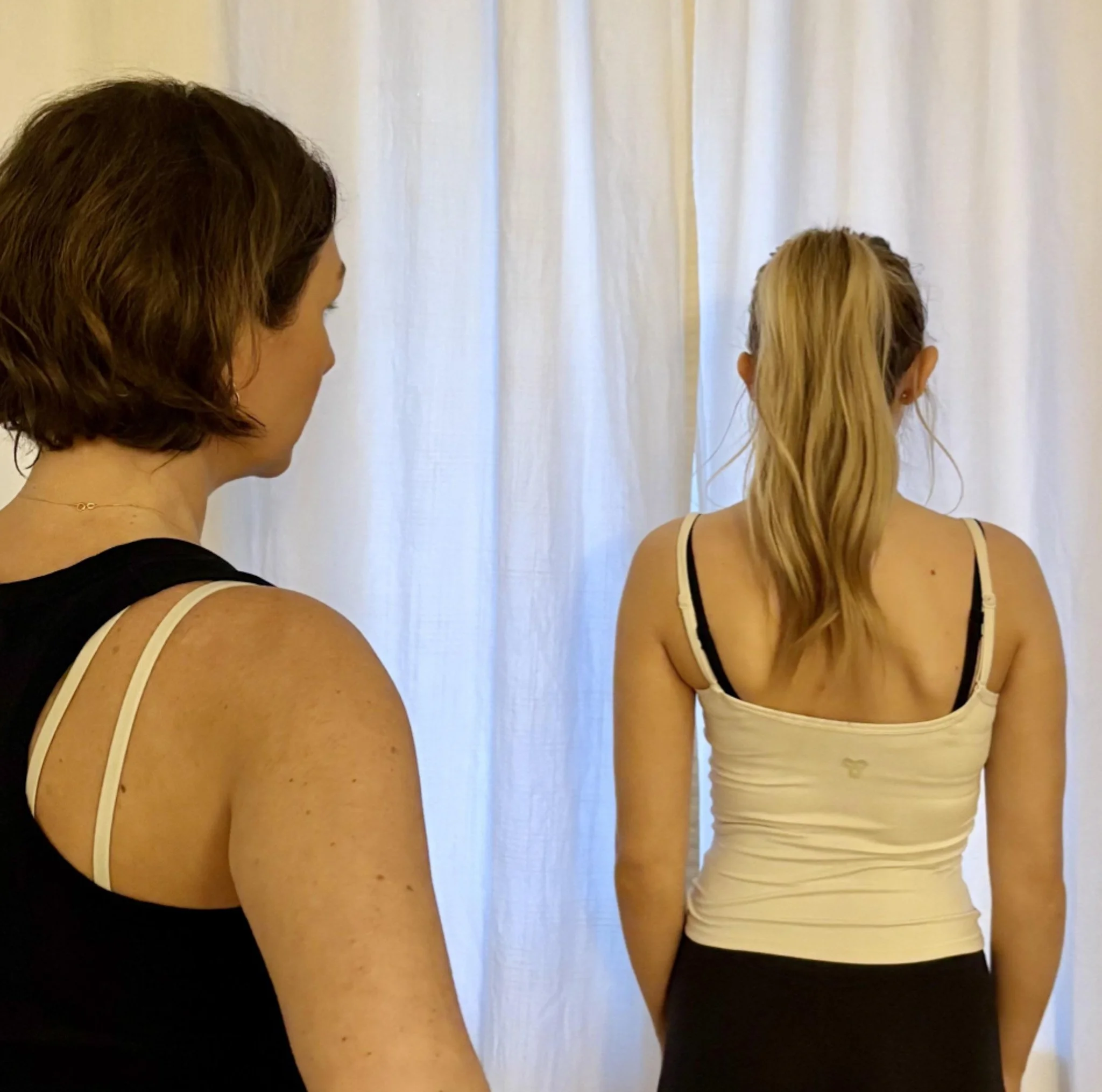 A woman with wavy brown hair and a woman with straight blonde hair, both wearing tank tops, are standing in front of a white curtain, facing away from the camera.