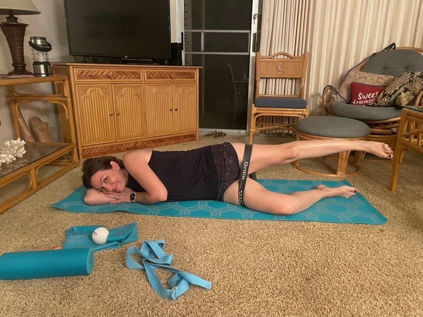A woman lying on her side on an exercise mat in a living room, with one leg raised and bent, performing a side leg raise exercise. She is smiling and resting her head on her hand, dressed in workout clothes.