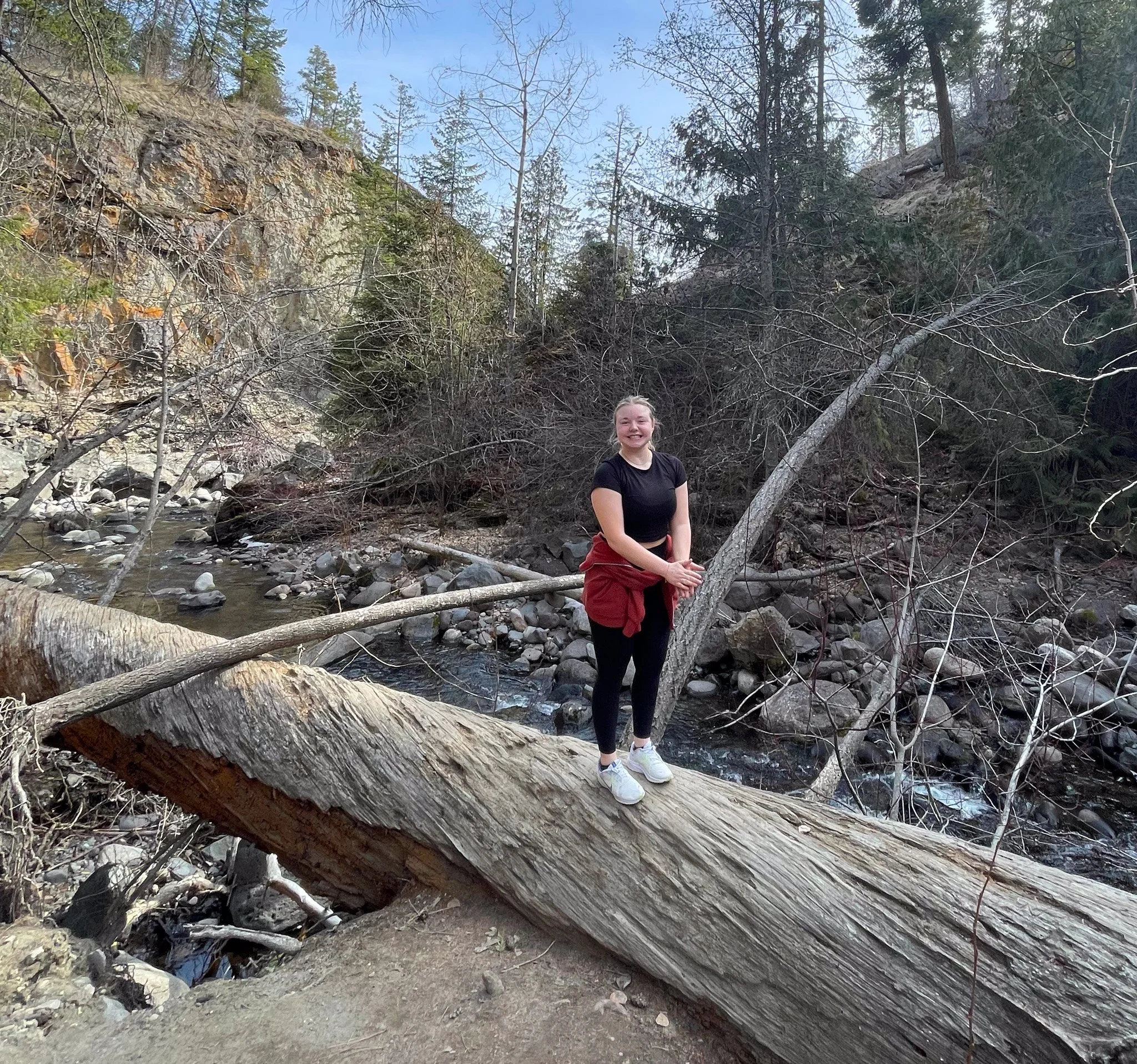 A young woman with a smiling face standing outdoors near a small creek in a wooded area with trees and rocks, with fallen logs and branches nearby.