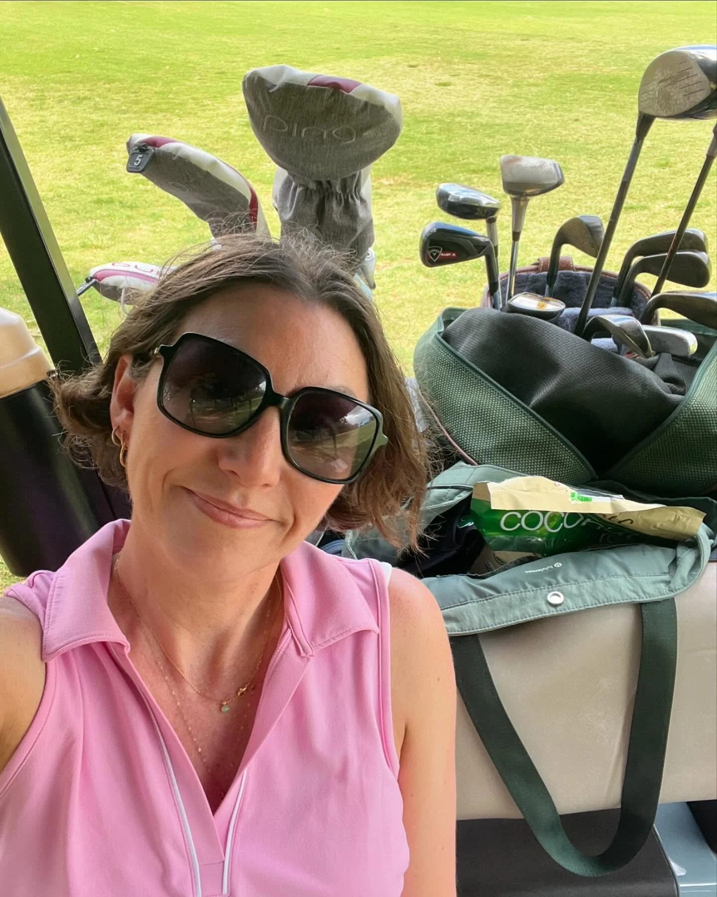 A woman wearing sunglasses and a pink sleeveless top taking a selfie inside a golf cart. The background shows a golf course with golf clubs and a bag behind her.