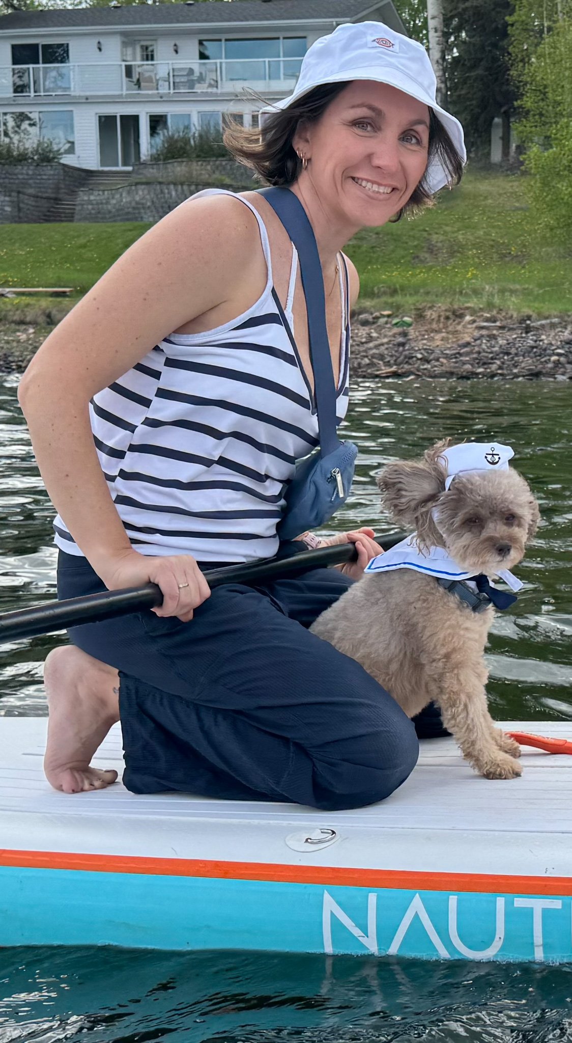 A smiling woman with short dark hair wearing a white sun hat and a striped tank top is kneeling on a paddleboard with a small dog dressed in a sailor outfit. They are on a body of water with a house and trees in the background.