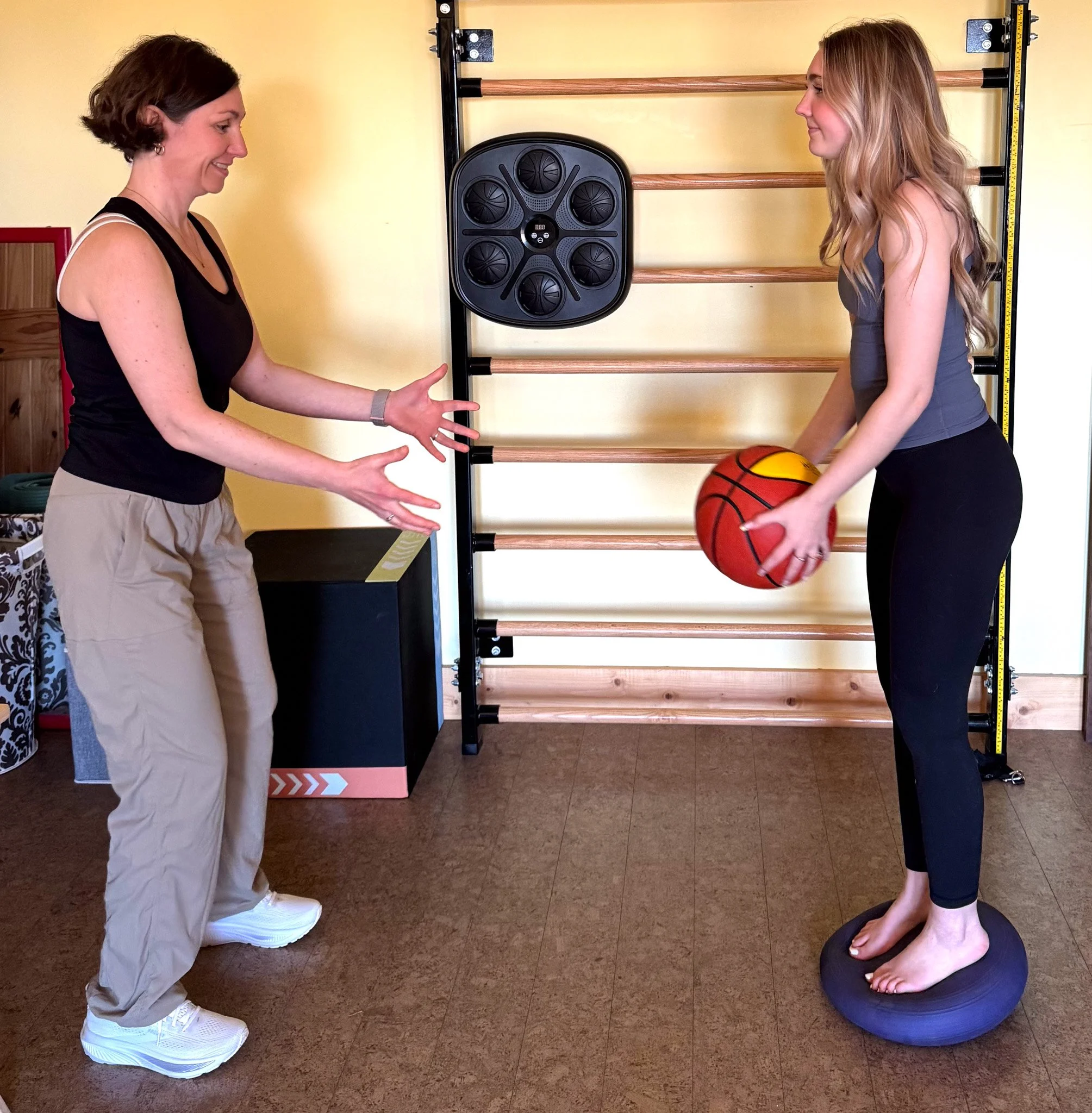 Two women in a fitness setting, one standing with arms extended and the other holding a basketball, with fitness equipment including a wall-mounted ladder and a multi-functional training pad in the background.