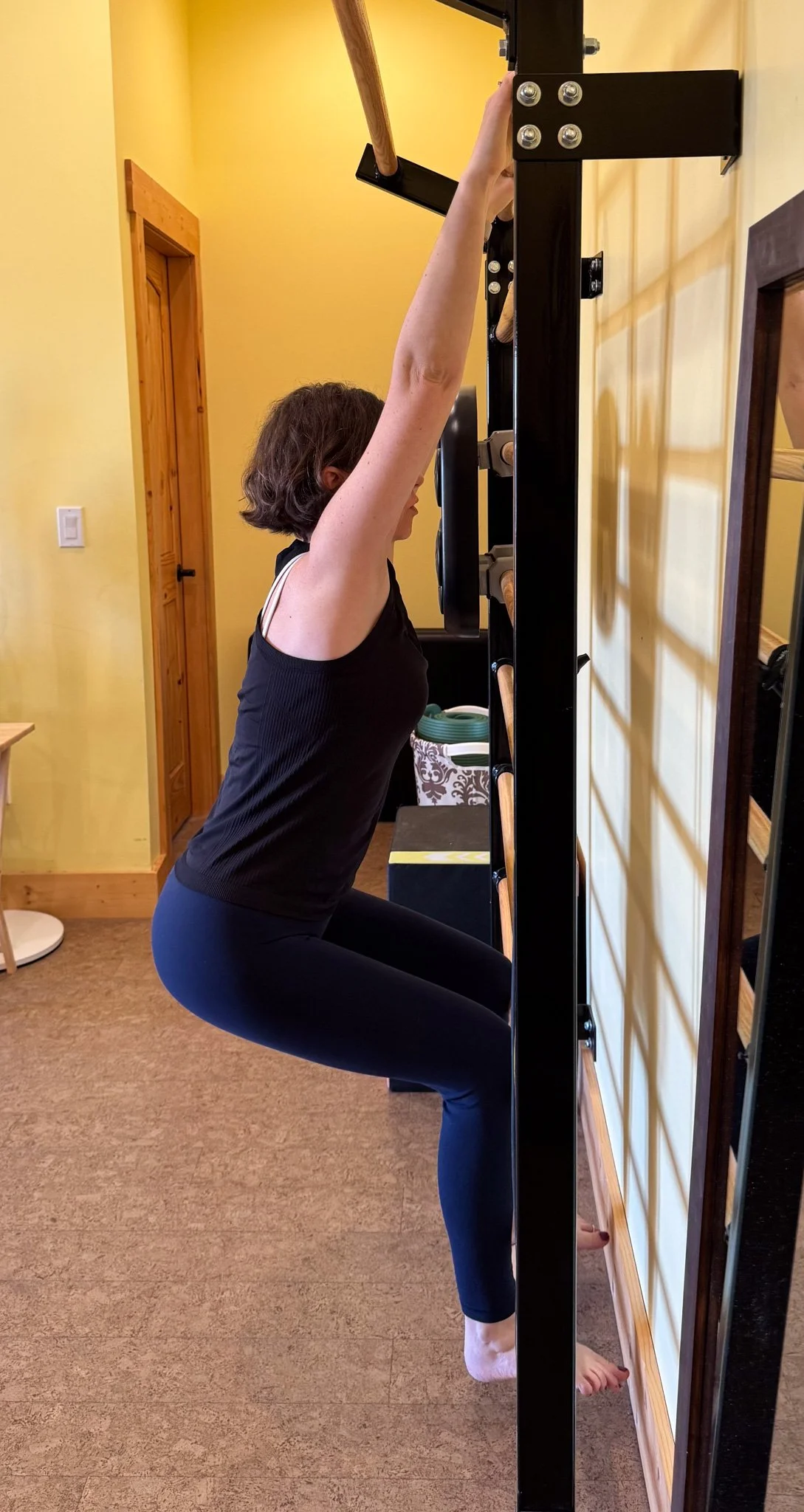 A woman performing a squat exercise using a pull-up bar mounted on a wood and metal frame, in a room with yellow walls and wooden accents.
