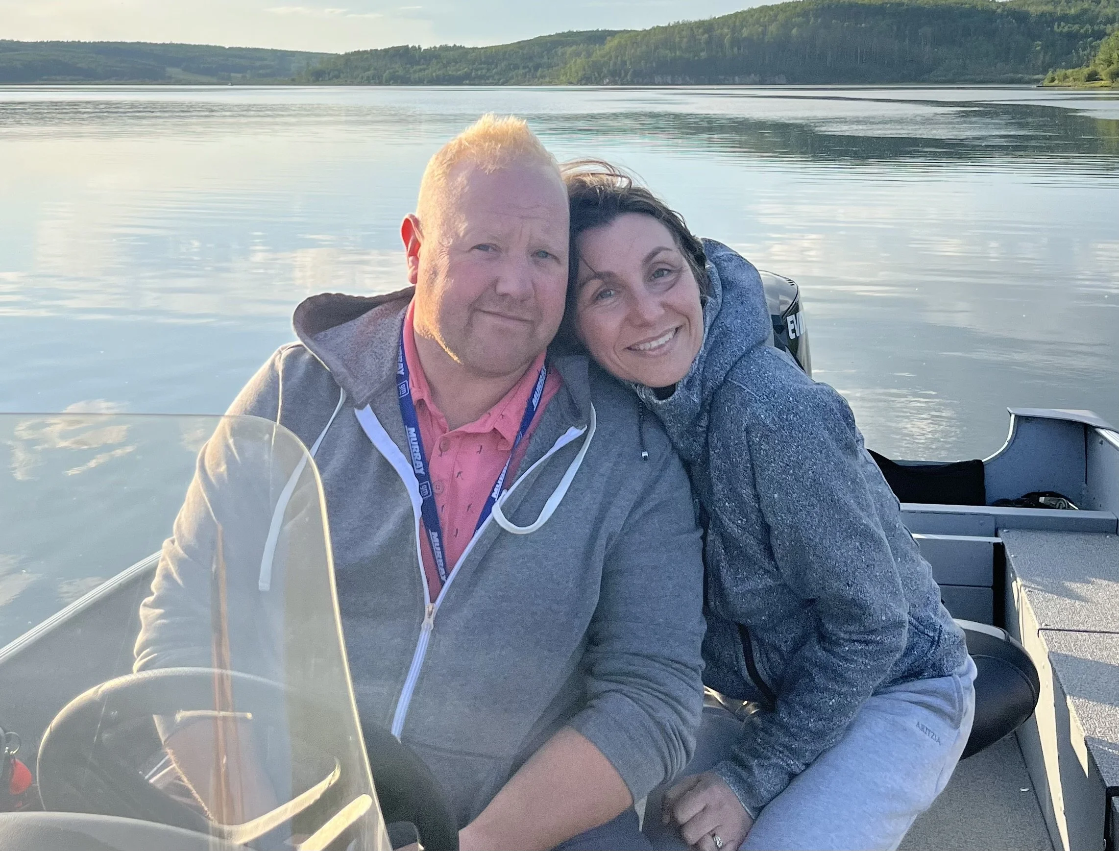 A smiling man and woman are sitting closely together on a boat, with a lake and green hills in the background during daytime.