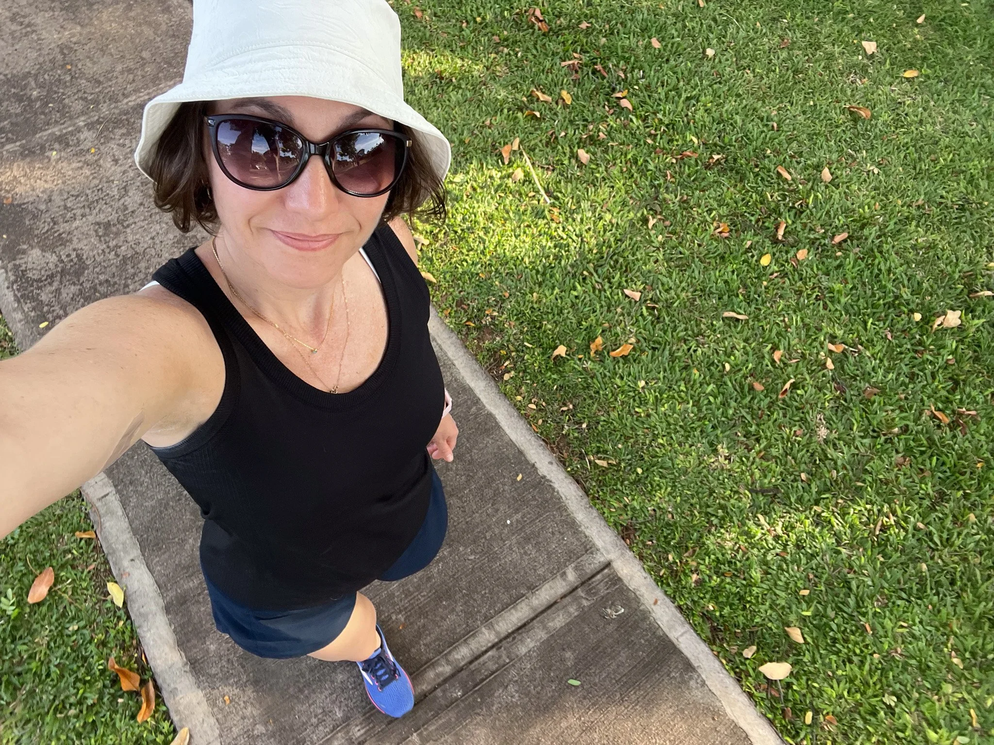 A woman taking a selfie outdoors, wearing a white bucket hat, large sunglasses, a black tank top, and dark shorts, standing on a sidewalk next to a grassy area with fallen leaves.