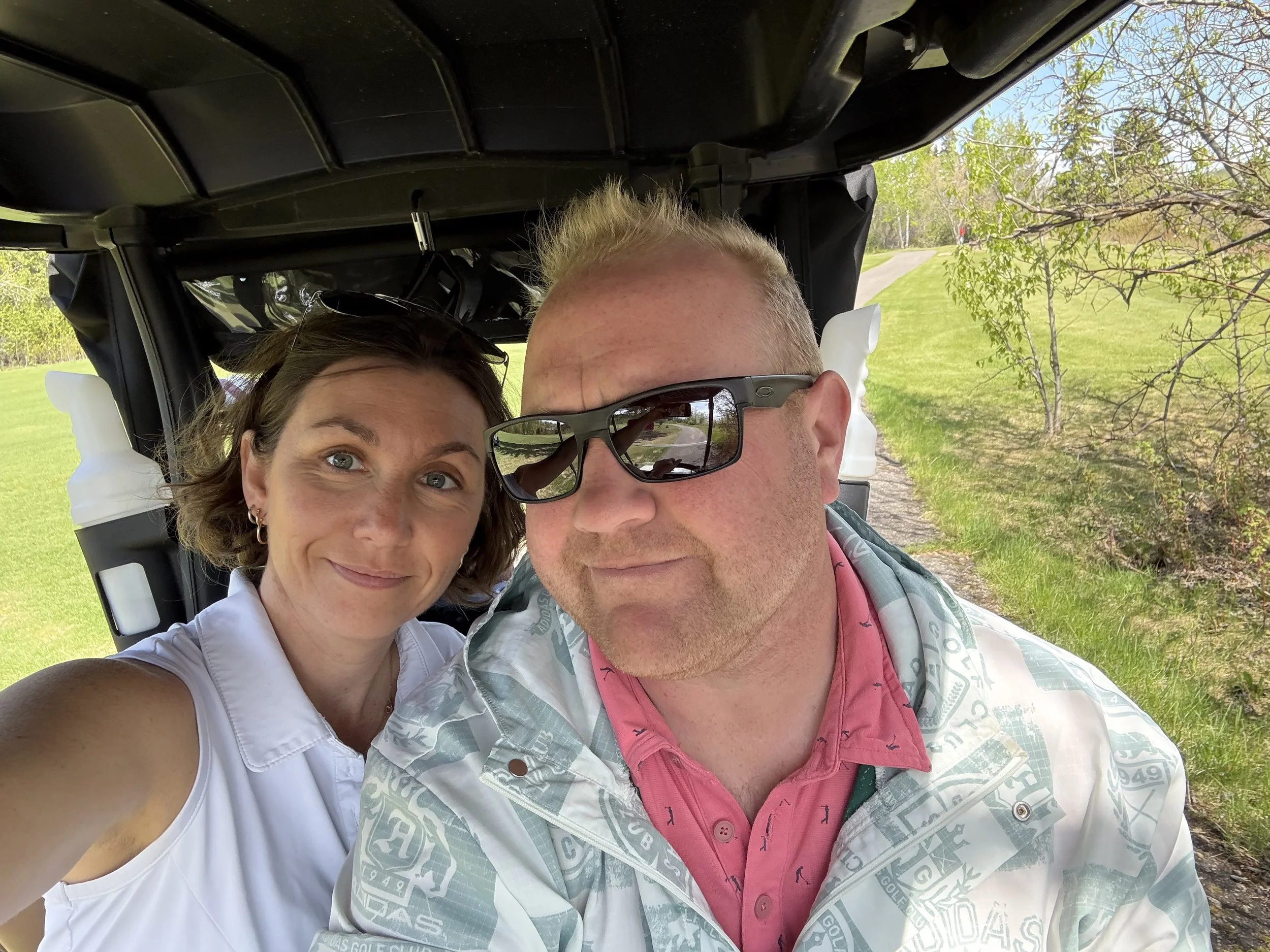 A man and woman taking a selfie while riding in a golf cart outside on a sunny day, with green grass, trees, and a path in the background.
