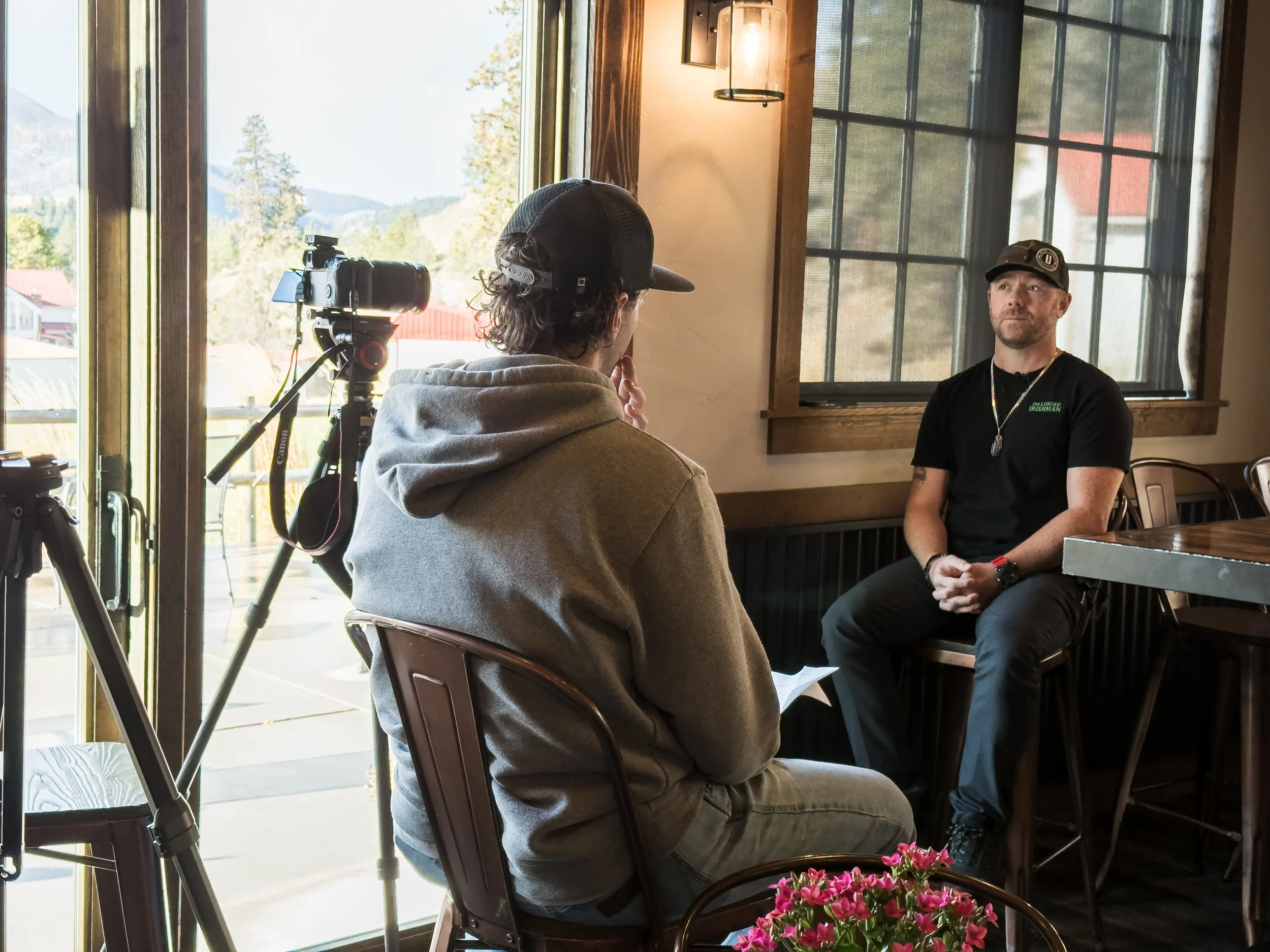 An indoor interview scene with two men, one facing away with a hoodie and cap, and the other in black clothing, sitting near large windows showing outdoors with trees and mountains.