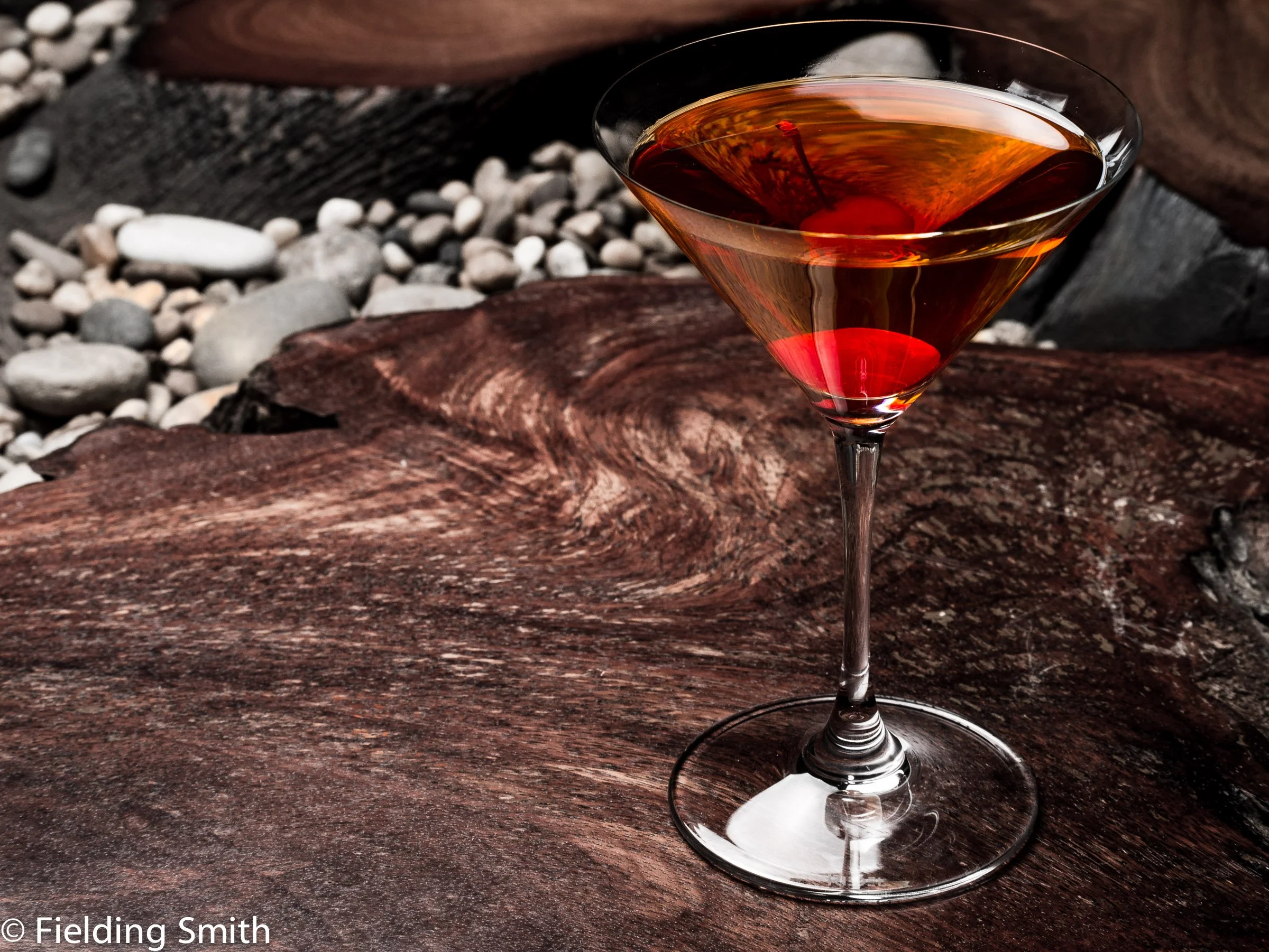 A martini glass filled with an amber-colored cocktail, garnished with a cherry at the bottom, placed on a textured wooden surface outdoors.