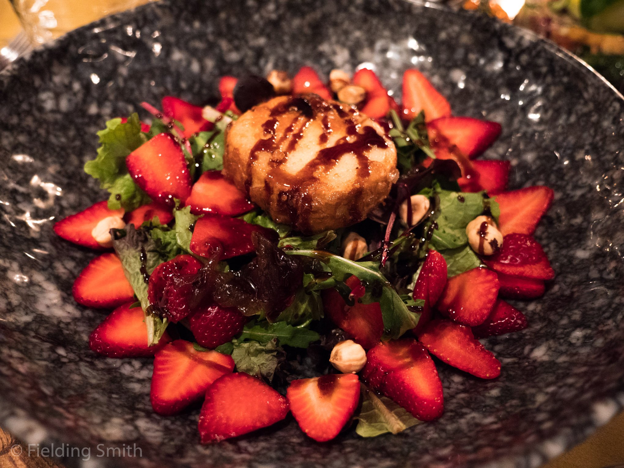 A salad with sliced strawberries, mixed greens, nuts, and a fried appetizer drizzled with balsamic glaze in a dark, textured bowl.