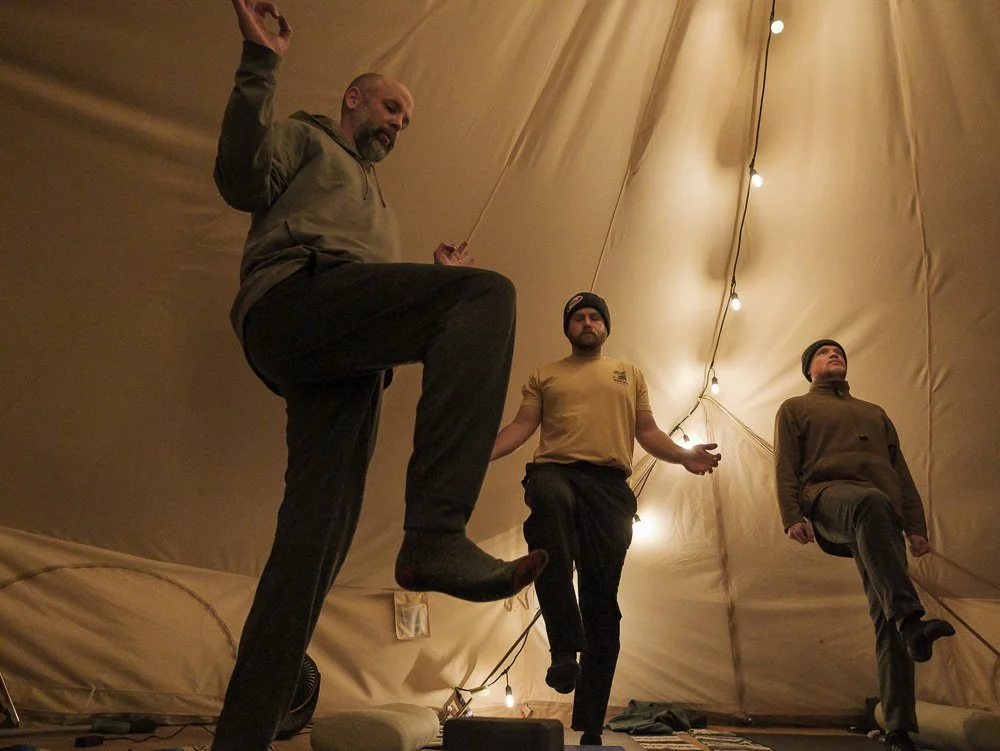 Three men practicing yoga inside a large tent lit by string lights, each balancing on one leg with their eyes closed.