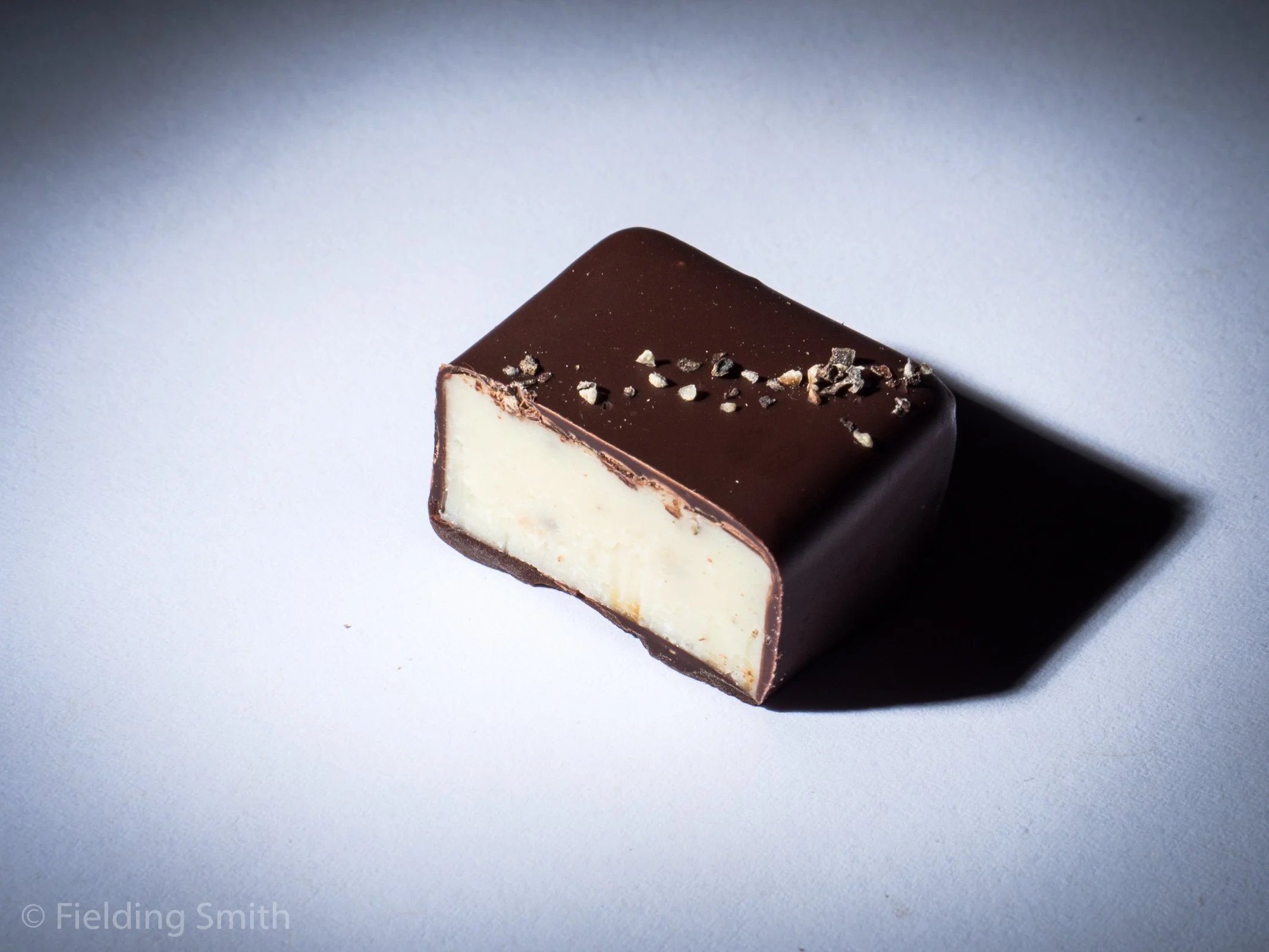 A square chocolate-covered confection with a white filling, garnished with small pieces of chocolate or nuts on top, resting on a white surface.