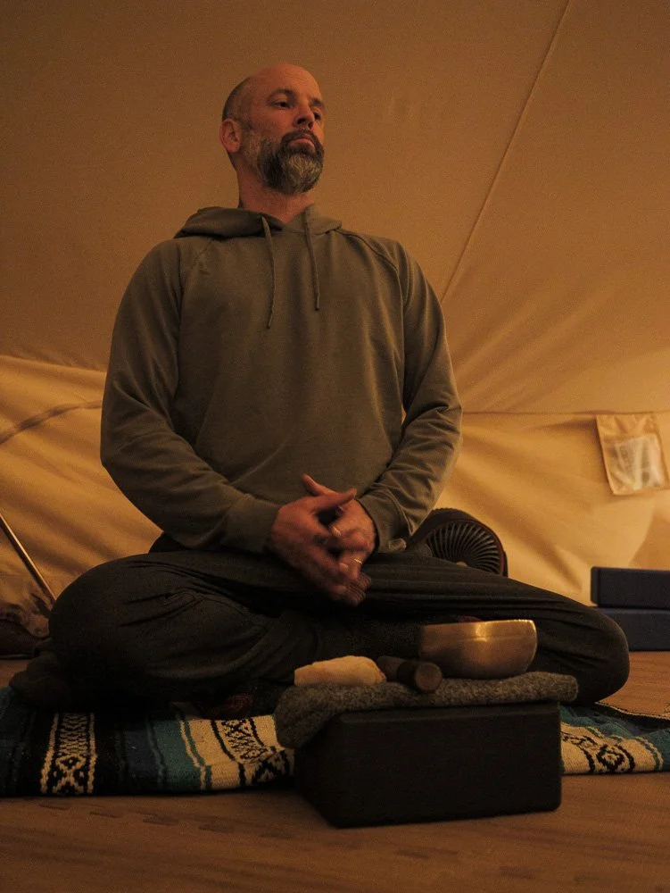 A man sitting cross-legged on a blanket inside a tent, with a singing bowl and other objects in front of him, possibly meditating or practicing mindfulness.