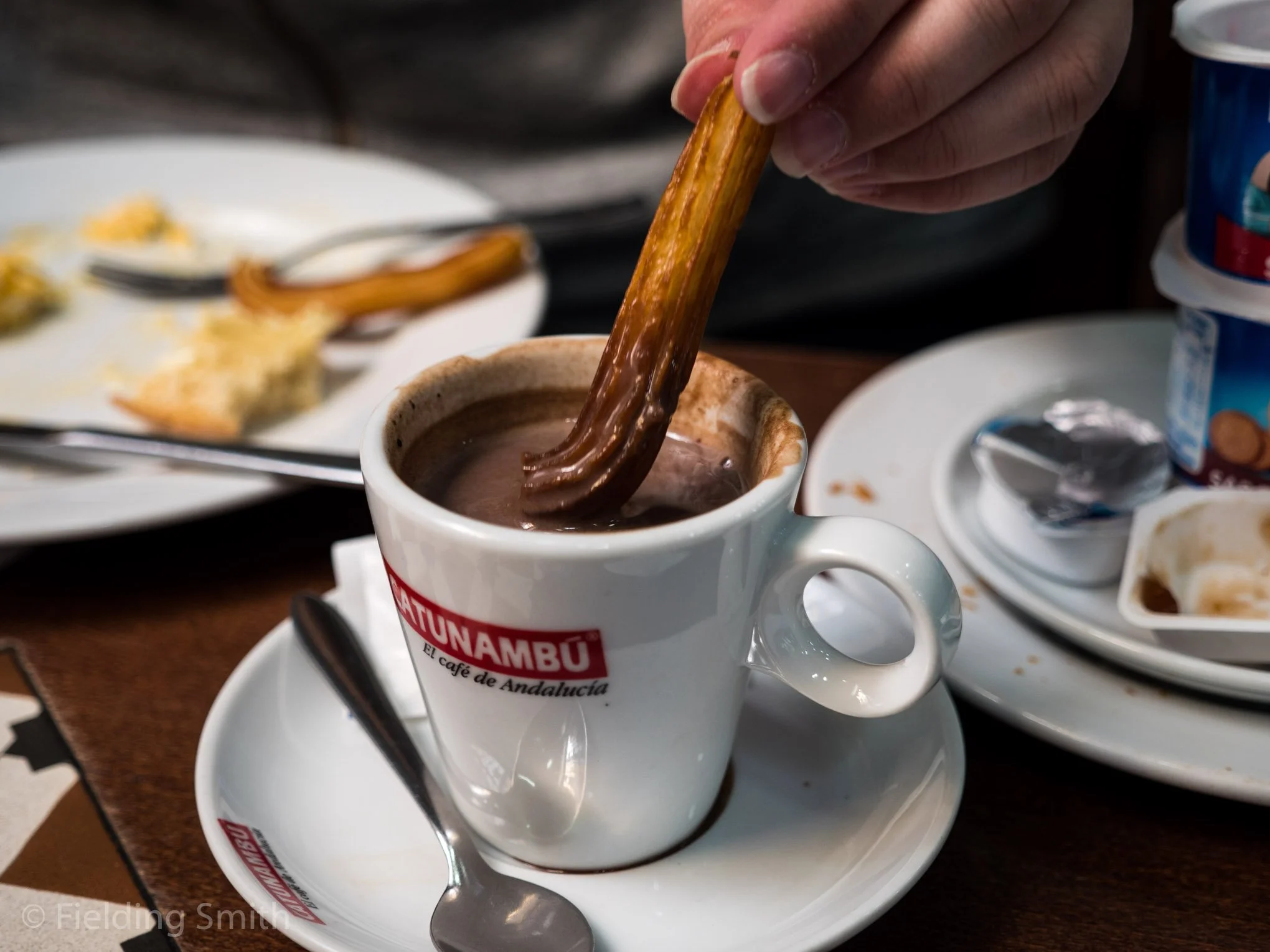 A person dunking a churro into a cup of hot chocolate on a table, with a plate of food and yogurt containers in the background.