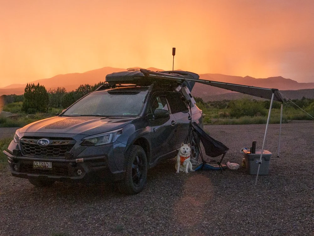A black Subaru SUV parked on a gravel surface during sunset with camping gear, a dog sitting beside it, and mountains in the background.