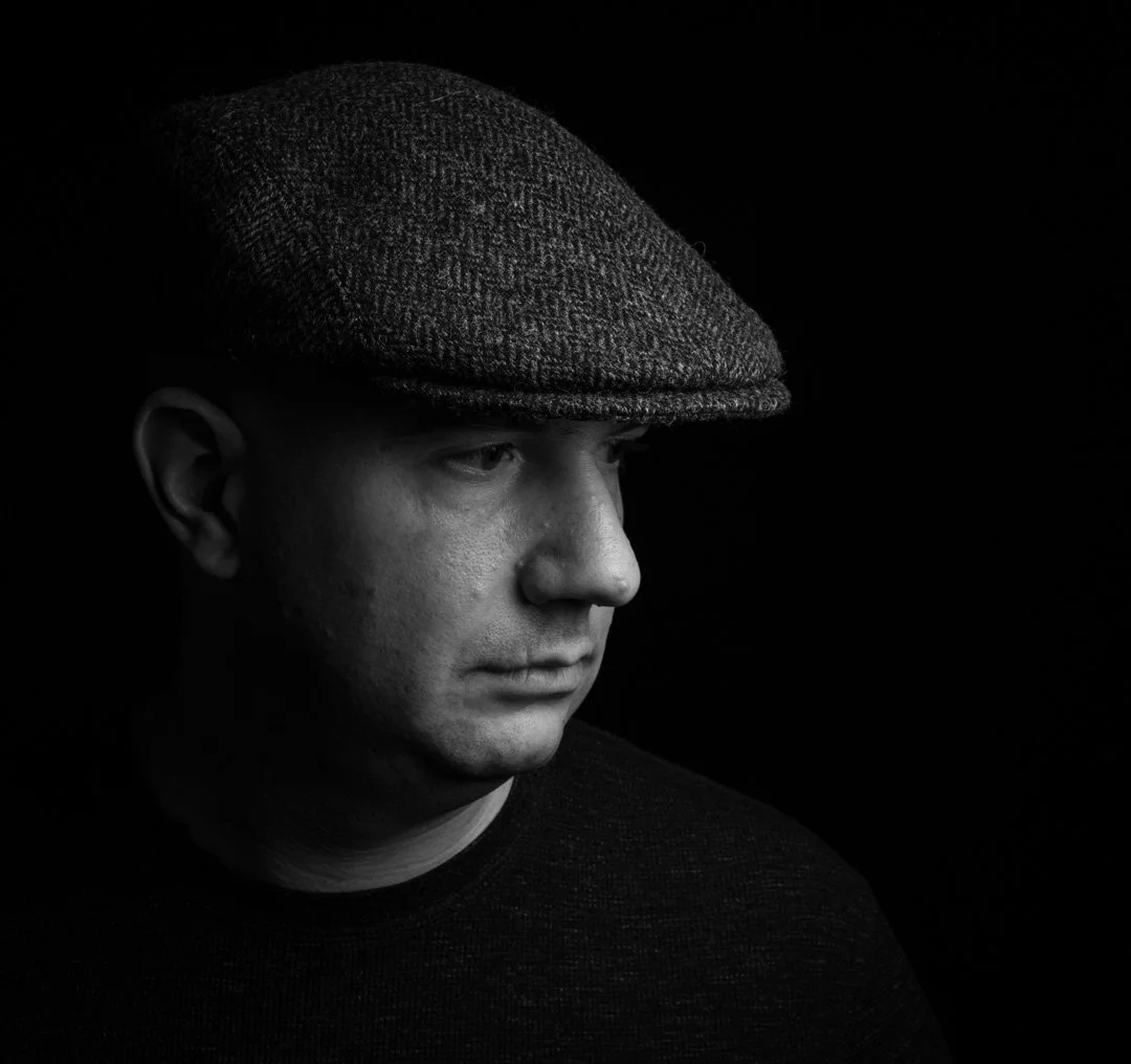 Black and white portrait of a man wearing a flat cap, facing left, with a serious expression, against a dark background.
