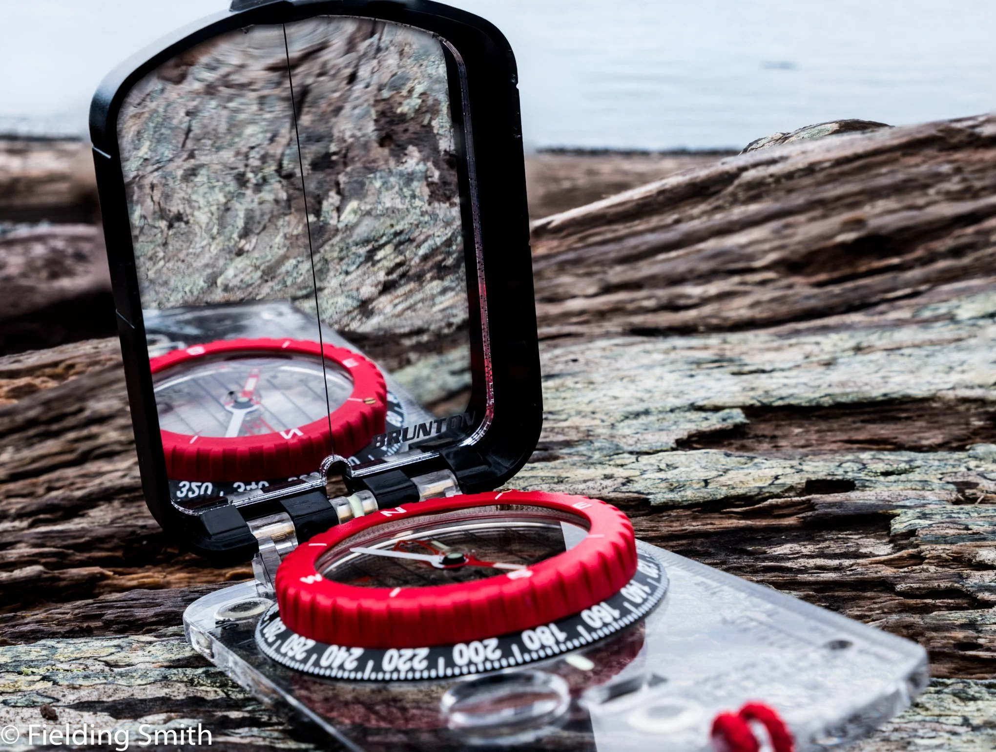 A compass with a red bezel, placed on a clear plastic case, is reflected in a mirror, showing a rocky landscape outside.