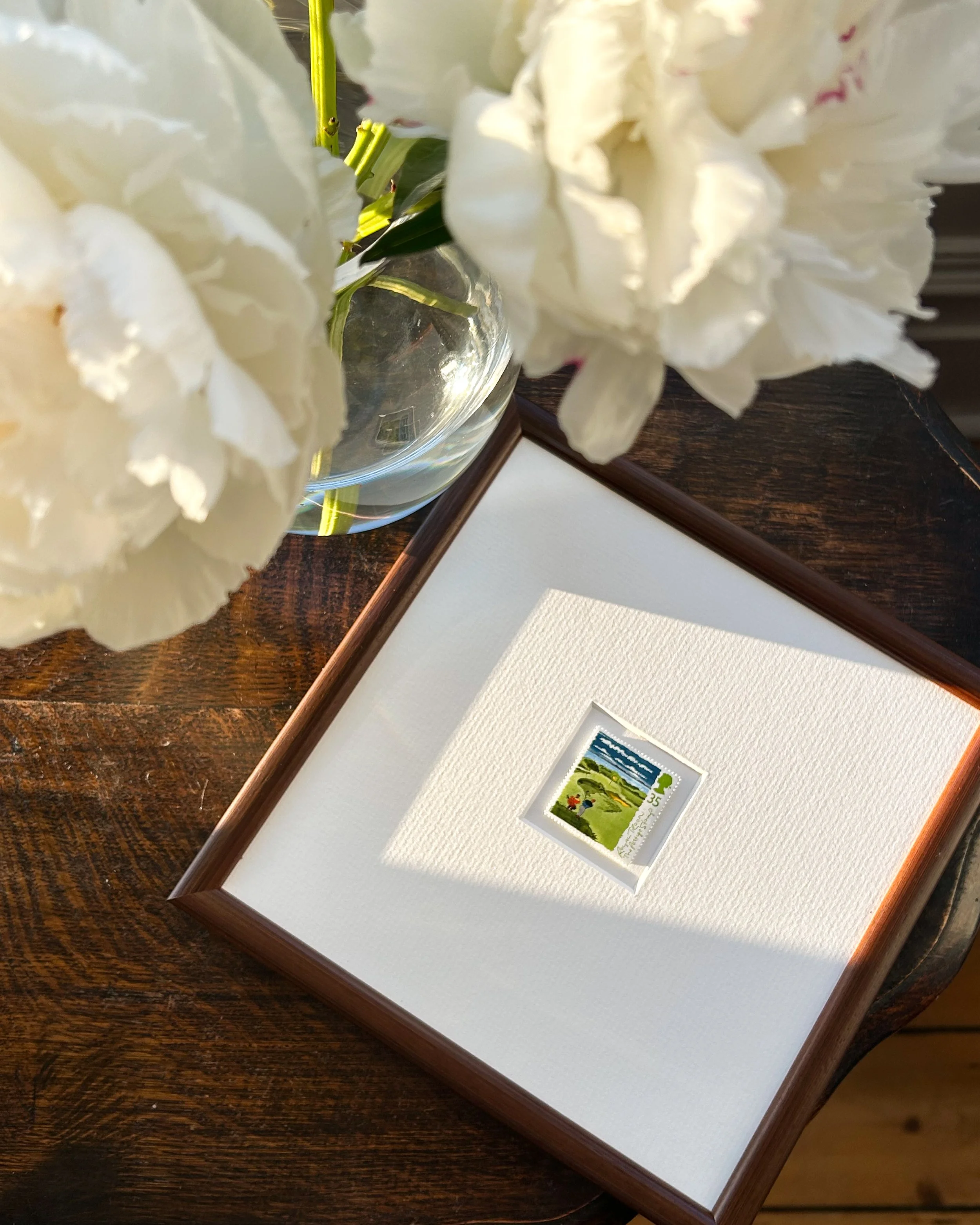 A framed stamp with an illustration of a golf course scene and small figures, framed within a white mount and brown wooden frame, resting on dark wooden table, with a bouquet of white flowers in a glass vase nearby.