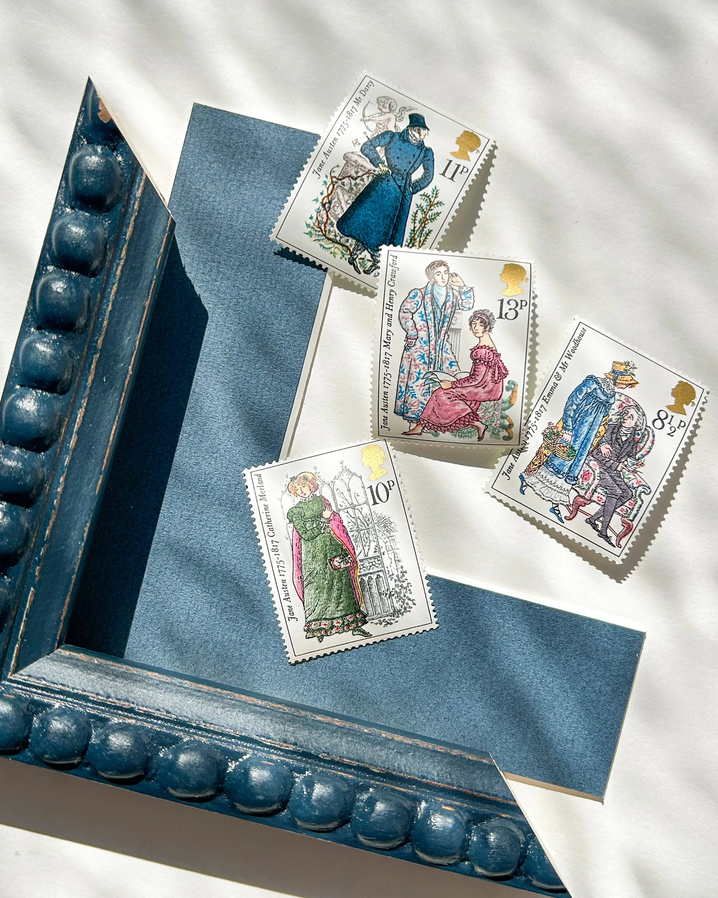 Close-up of vintage postage stamps featuring characters from Jane Austen's writings, with a blue mount board and a blue frame sample.