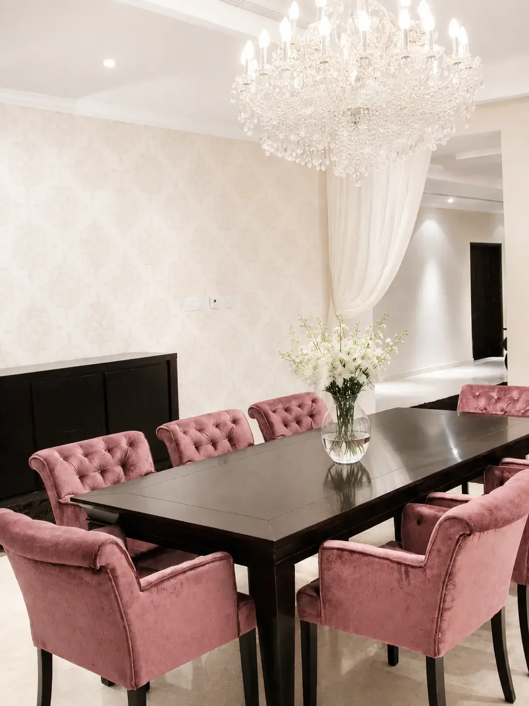 Elegant dining room with blush velvet chairs, crystal chandelier, dark wood table, and soft neutral interiors in a luxury villa.