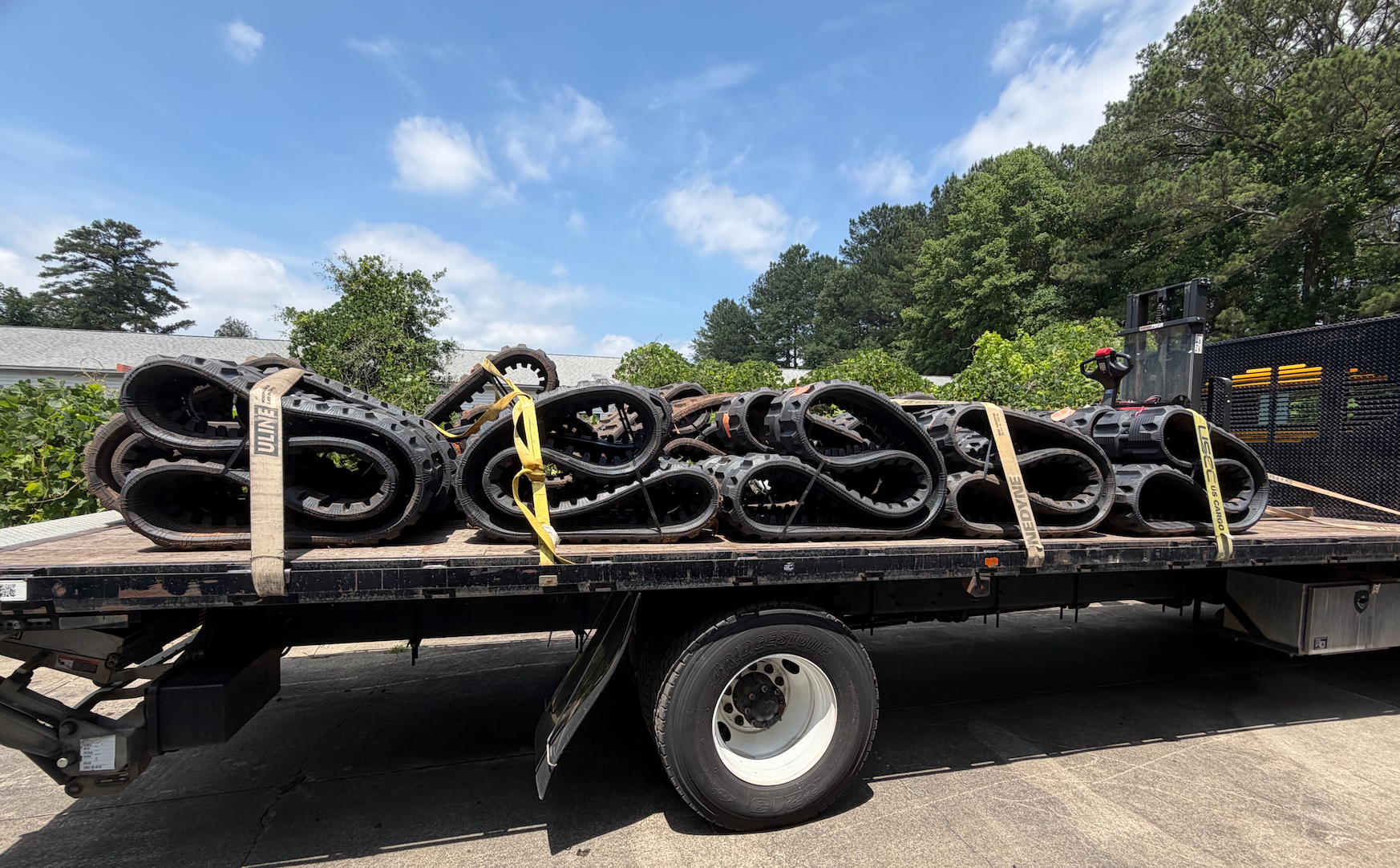 Flatbed truck carrying several black rubber tracks, possibly for construction equipment, secured with yellow straps, parked outdoors with trees and blue sky in the background.