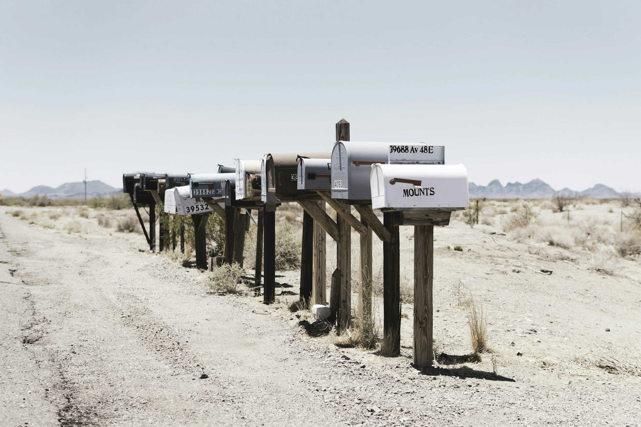 Multiple mailboxes mounted on wooden posts, situated in a desert landscape with dry bushes and mountains in the background.
