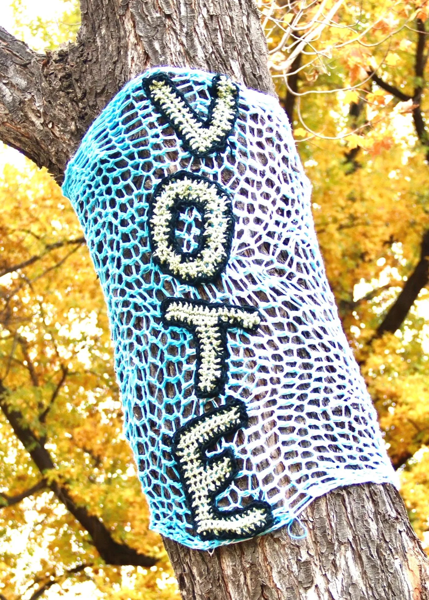 A navy blue and white crocheted mesh bag hanging on a tree trunk during autumn, with colorful fall leaves in the background.