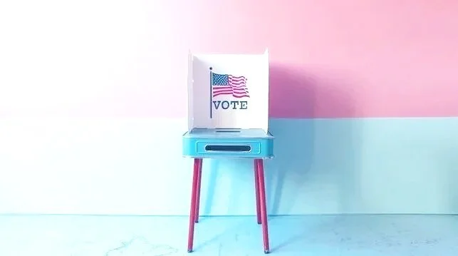 A voting booth with an American flag and the word 'VOTE' on it, placed on a small table with pink legs, against a pink and white background.